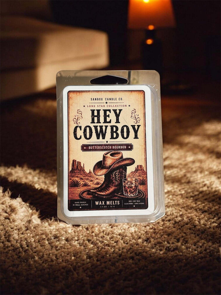 Hey Cowboy soy wax melt package featuring a cowboy boot and desert illustration, displayed on a rug beside a warm lamp — Sanbox Candle Co. Lone Star Collection