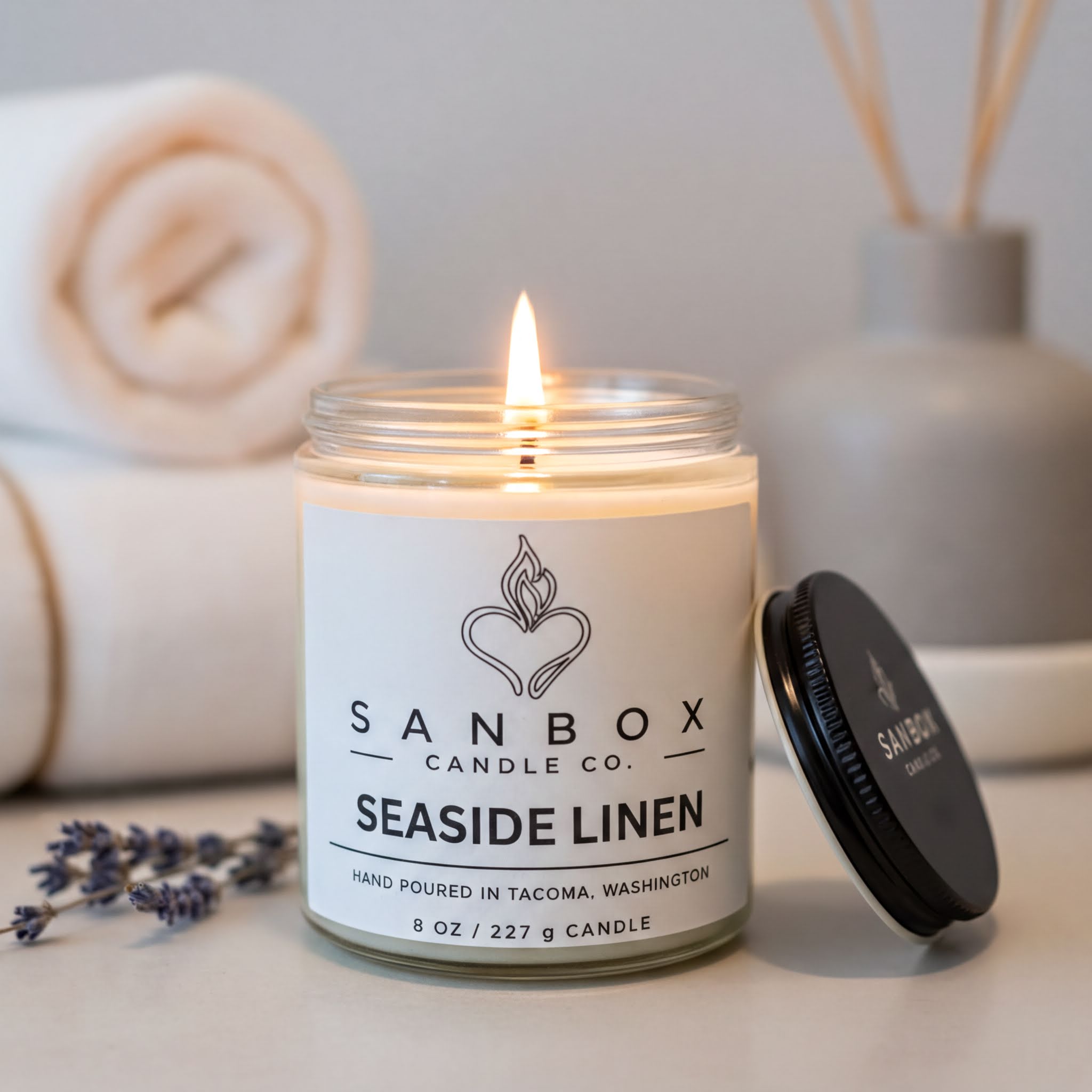 A lit candle in a jar labeled "SANBOX CANDLE CO. SEASIDE LINEN" sits on a table, with a rolled towel and a vase of sticks in the background.