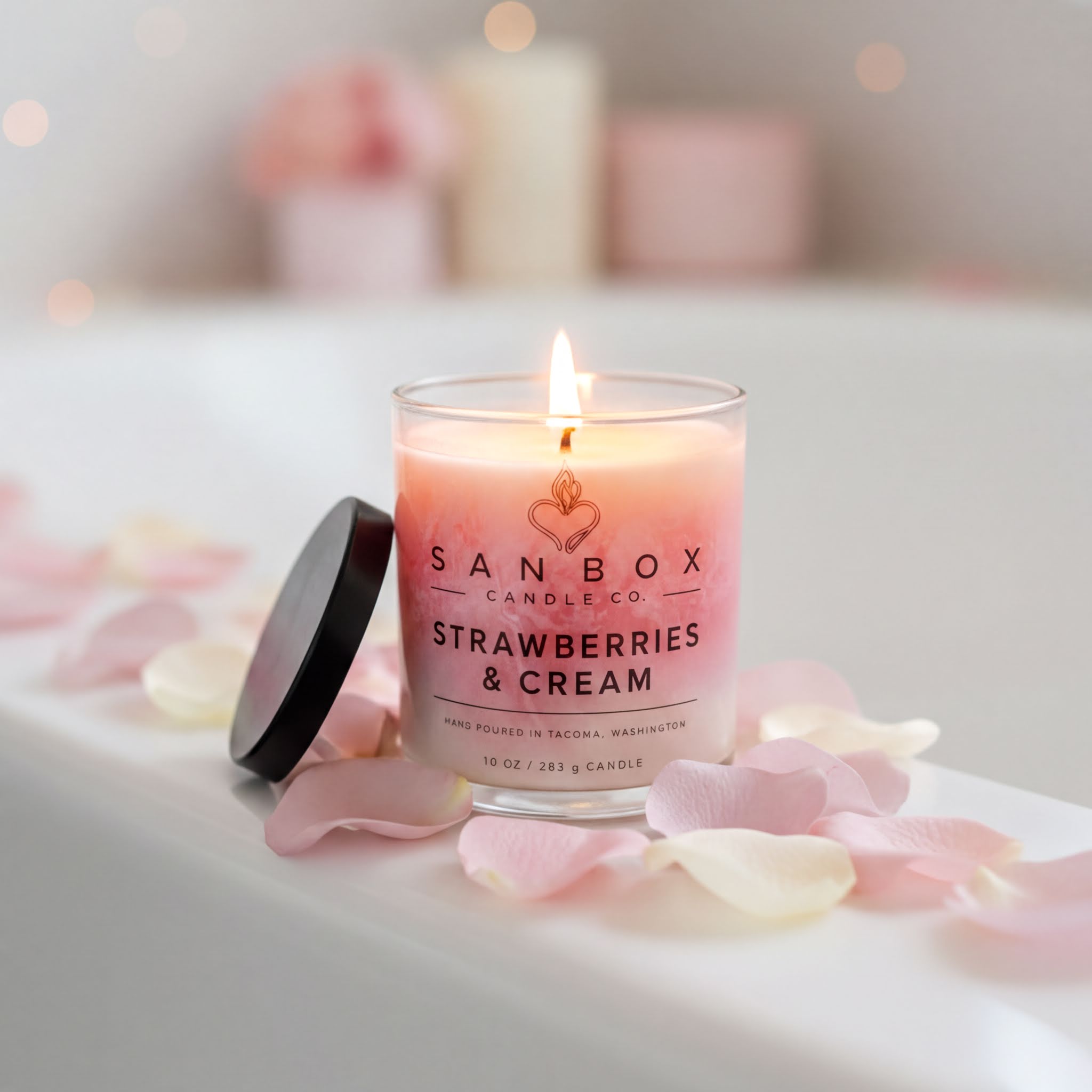 A lit candle in a glass container with a black lid, surrounded by pink rose petals, on a white surface.