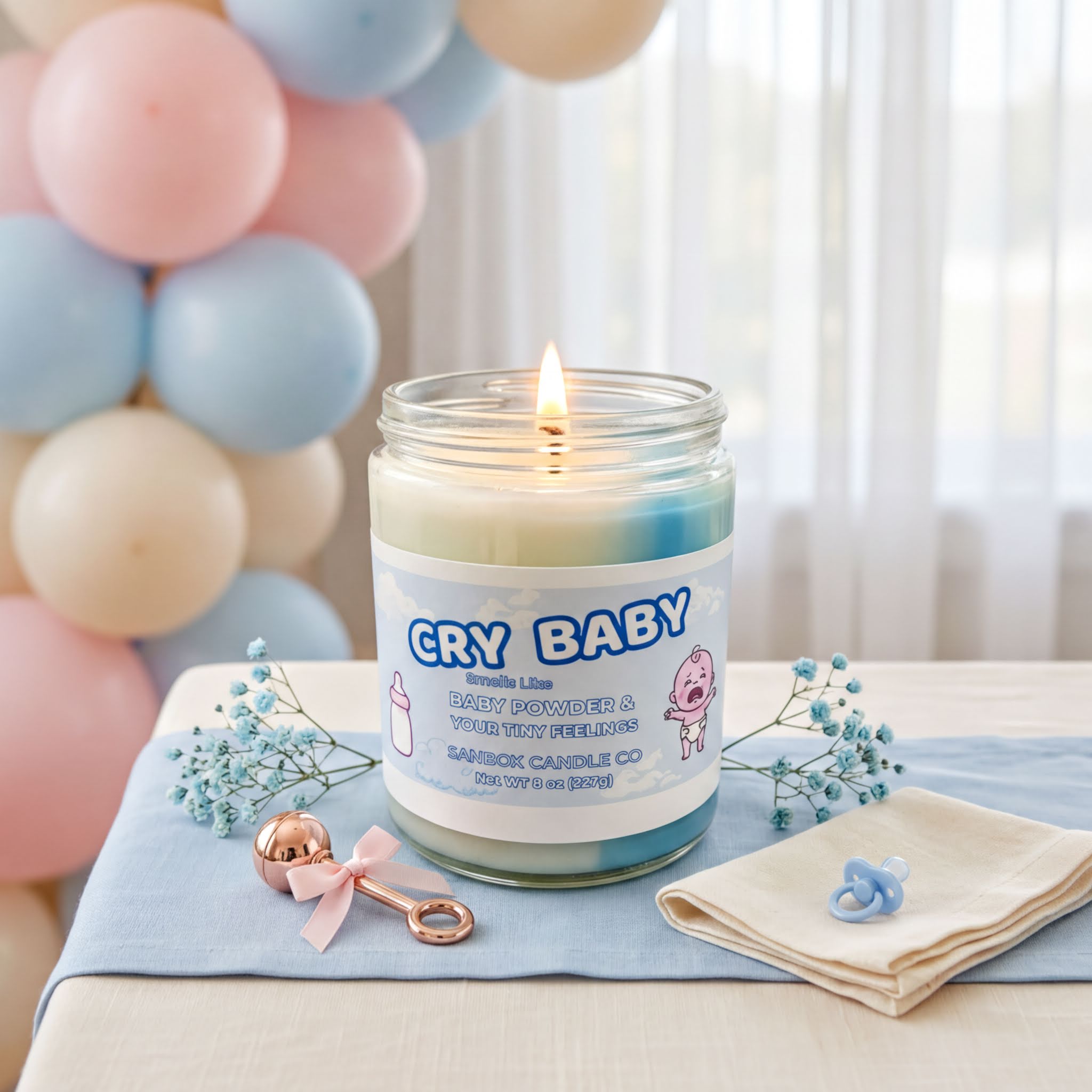 a candle labeled "CRY BABY" on a table, surrounded by baby-related items such as a pacifier, a baby bottle, and a blue napkin.
