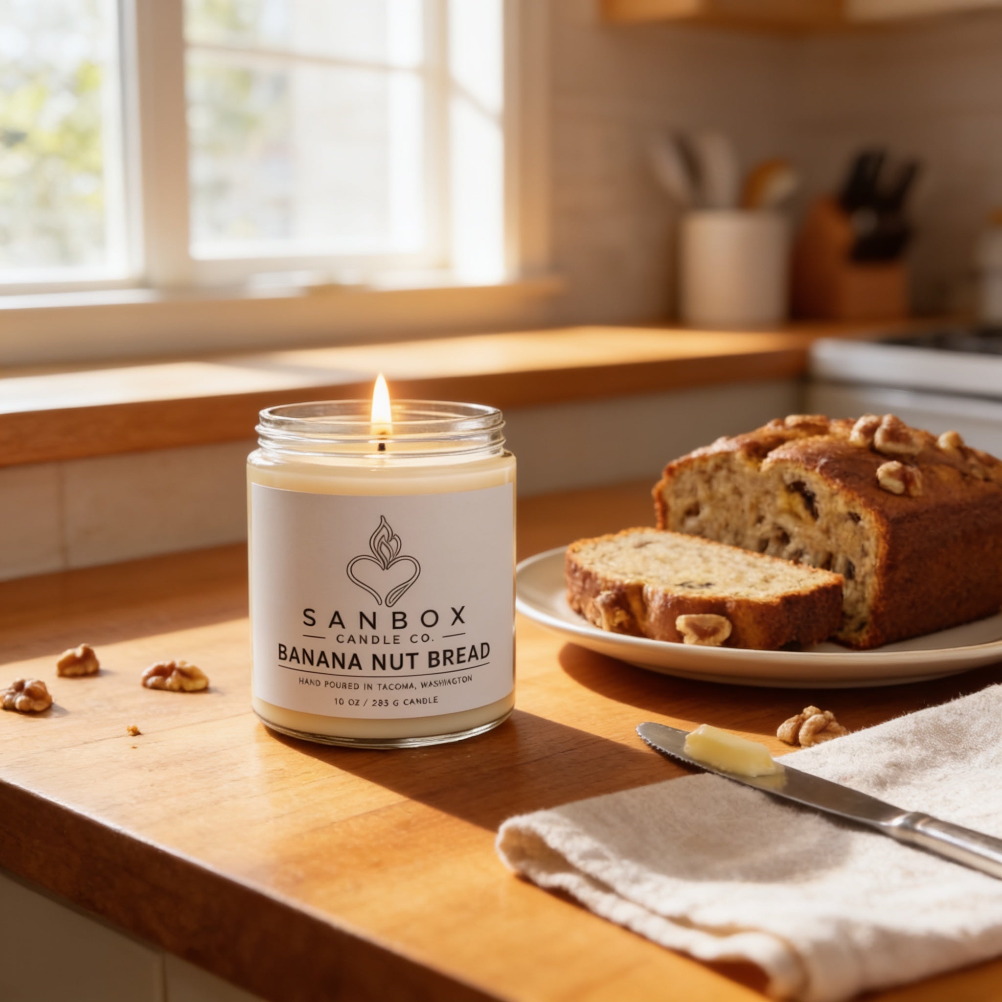 A candle labeled "Sanbox Candle of Banana Nut Bread" sits on a wooden counter, with a slice of banana nut bread and a knife nearby.