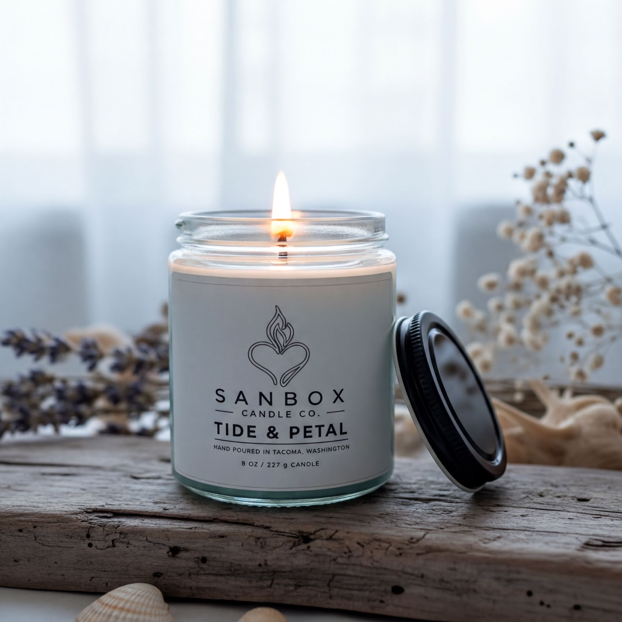A lit candle in a glass jar with a black lid, placed on a wooden surface, with some flowers and a plant in the background.