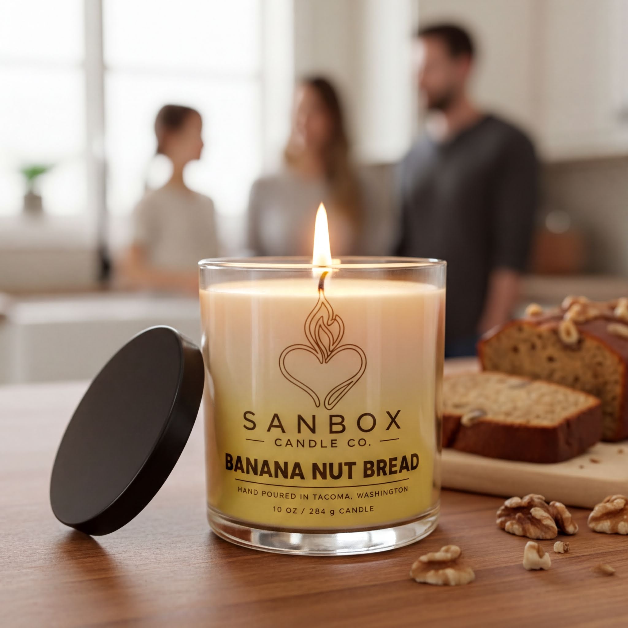 A lit candle in a glass container labeled "SANBOX BANANA NUT BREAD" sits on a wooden table, with a slice of banana nut bread and some walnuts nearby.
