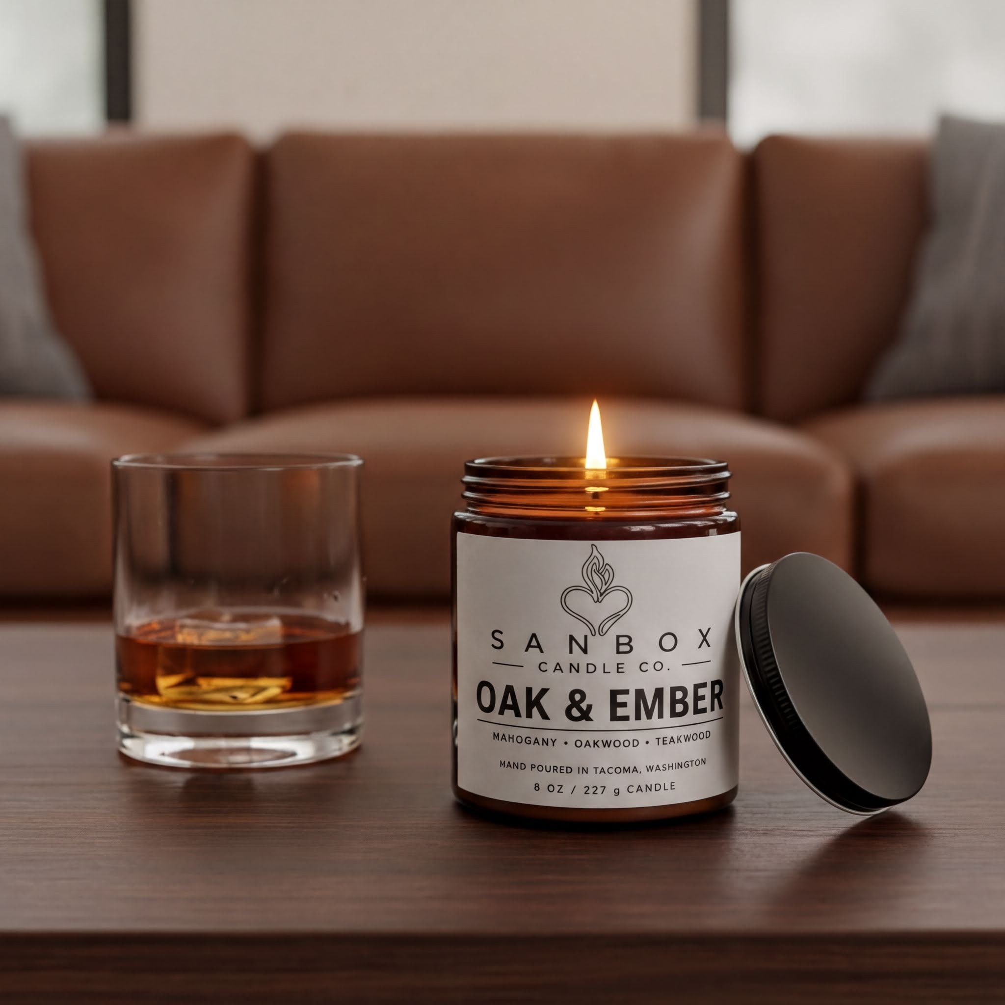 a candle labeled "SANBOX" and "OAK & EMBER" on a wooden table, with a glass of amber liquid next to it.