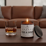 a candle labeled "SANBOX" and "OAK & EMBER" on a wooden table, with a glass of amber liquid next to it.