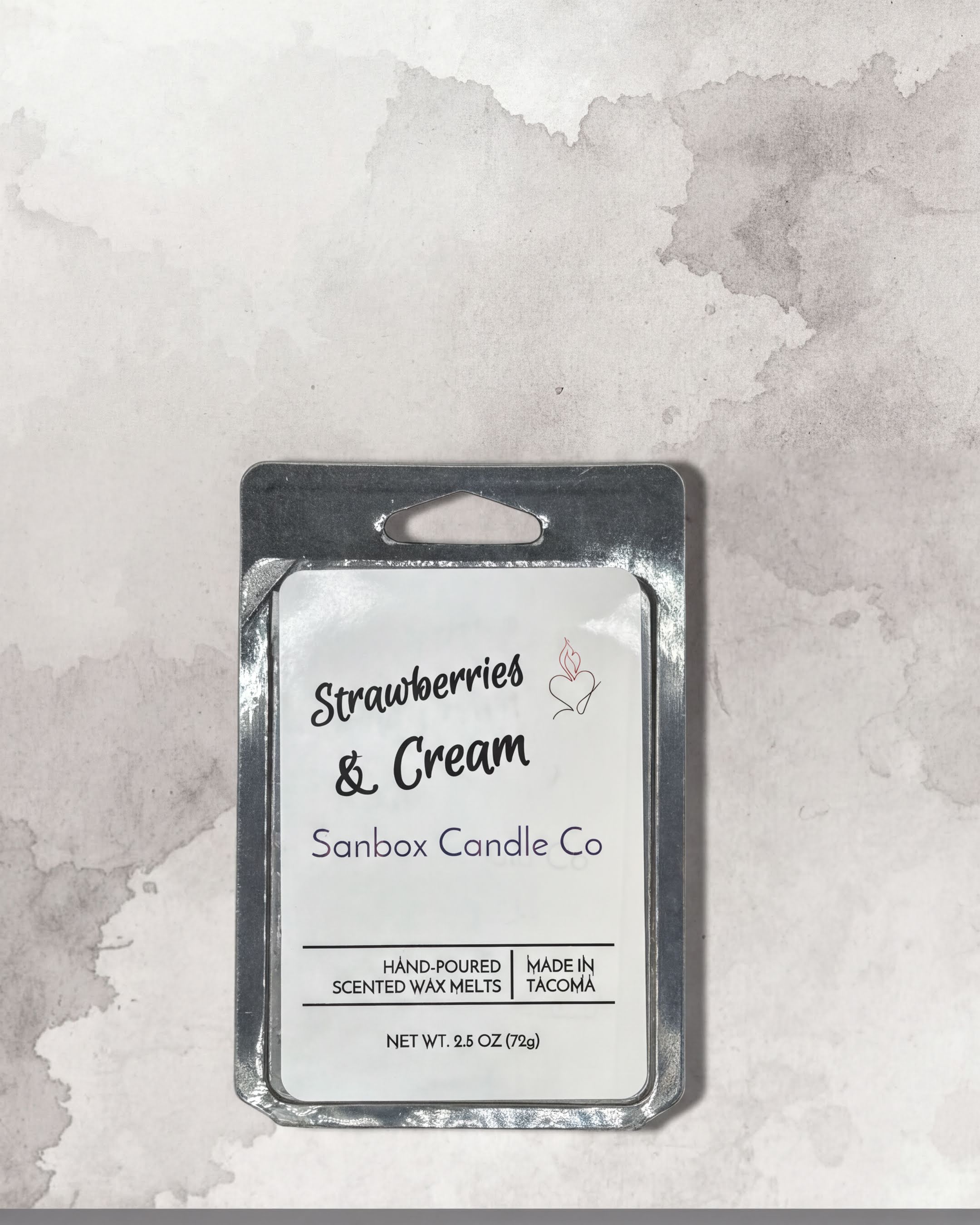 a rectangular metal container with a label that reads "Strawberries & Cream Sanbox Candle Co." The container is placed on a textured gray background.