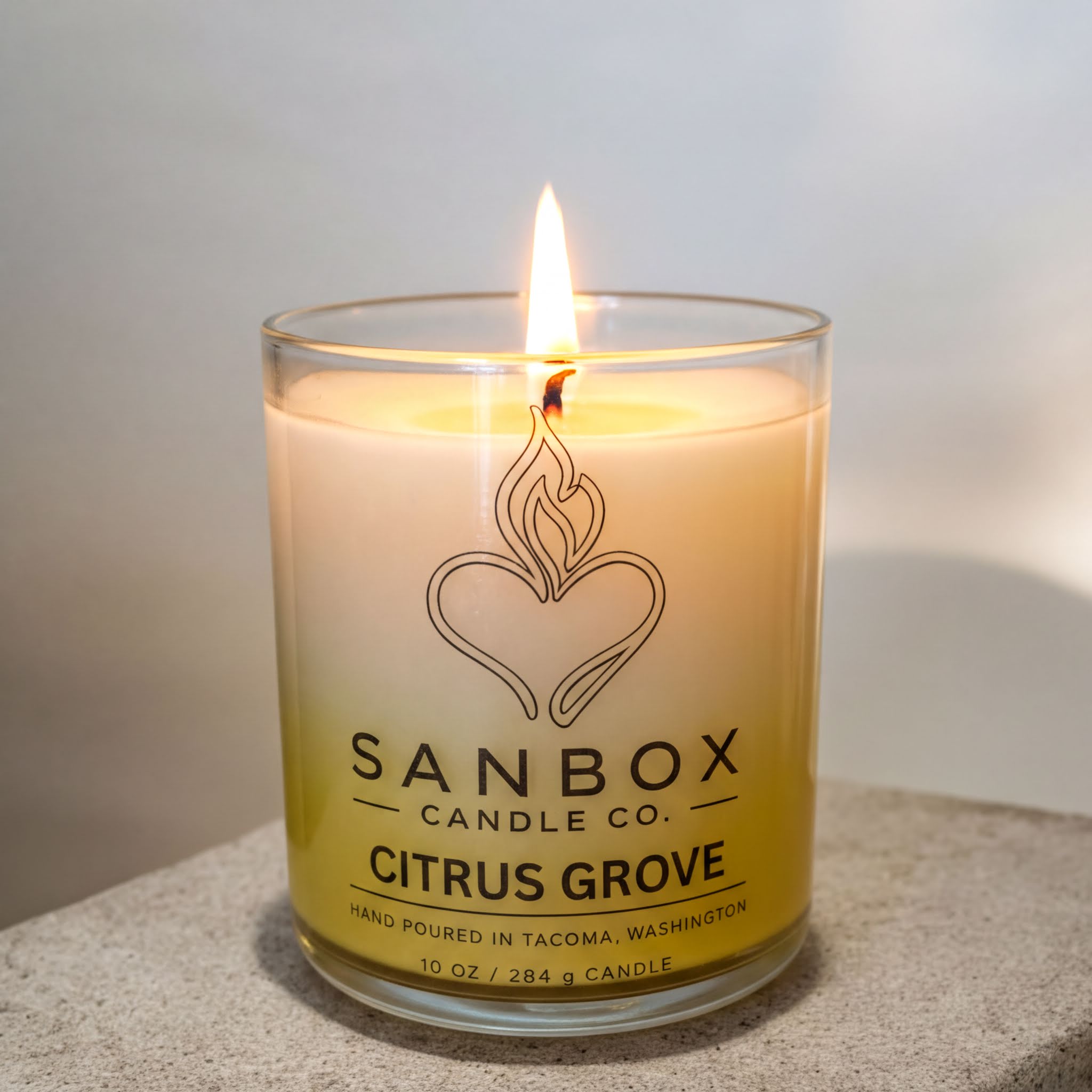 A lit candle in a glass container, with a label that reads "SANBOX CANDLE CO. CITRUS GROVE" and "HAND POURED IN TACOMA, WASHINGTON".