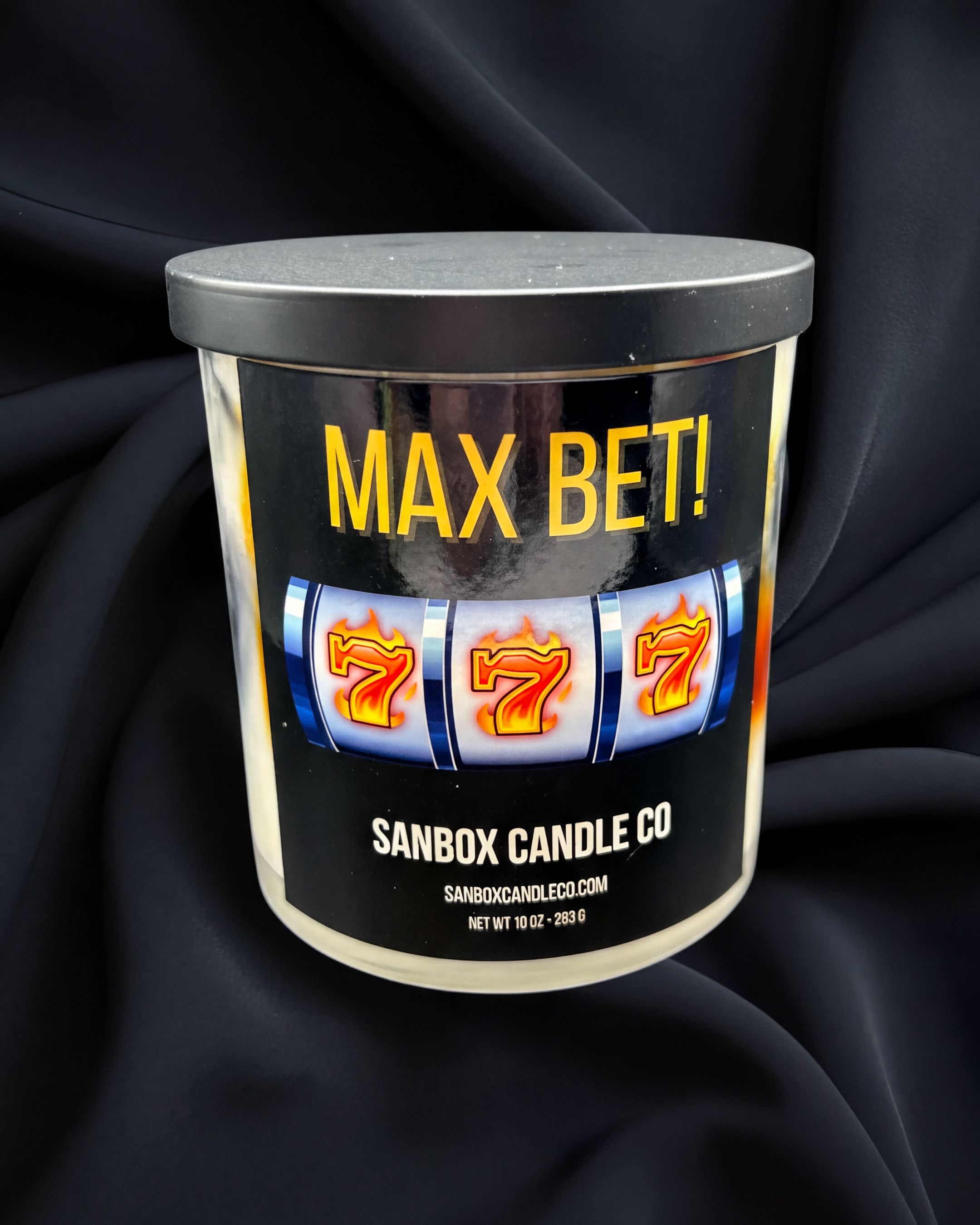 a cylindrical container with a black lid, placed on a black fabric background. The container has a label that reads "MAX BET!" and "SANBOX CANDLE CO".