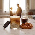 a candle labeled "SANBOX CASHMERE SPICE" on a kitchen counter, with a cup of coffee and a pretzel nearby.