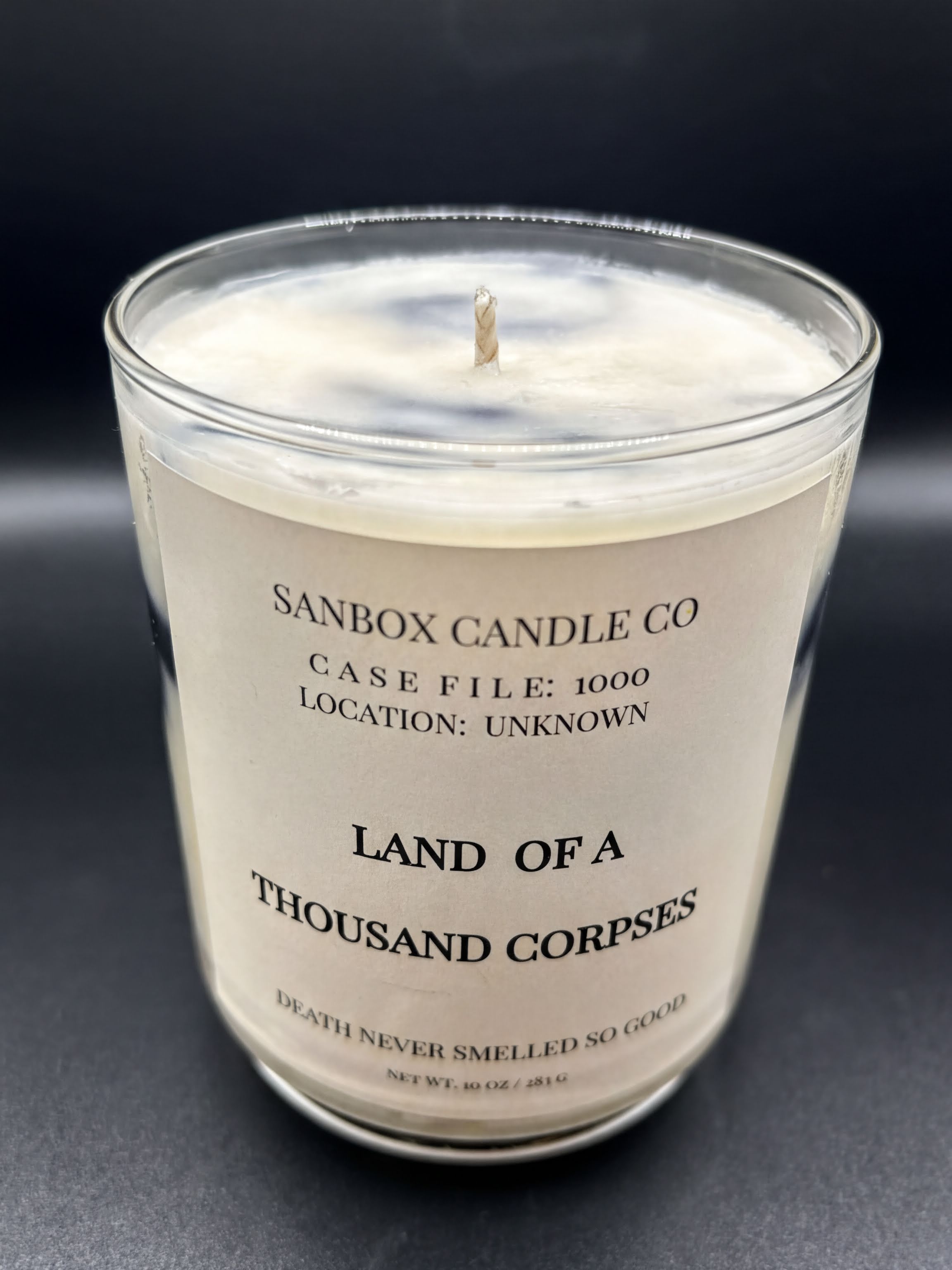 Scented candle in clear glass jar with Land of a Thousand Corpses label on black background.