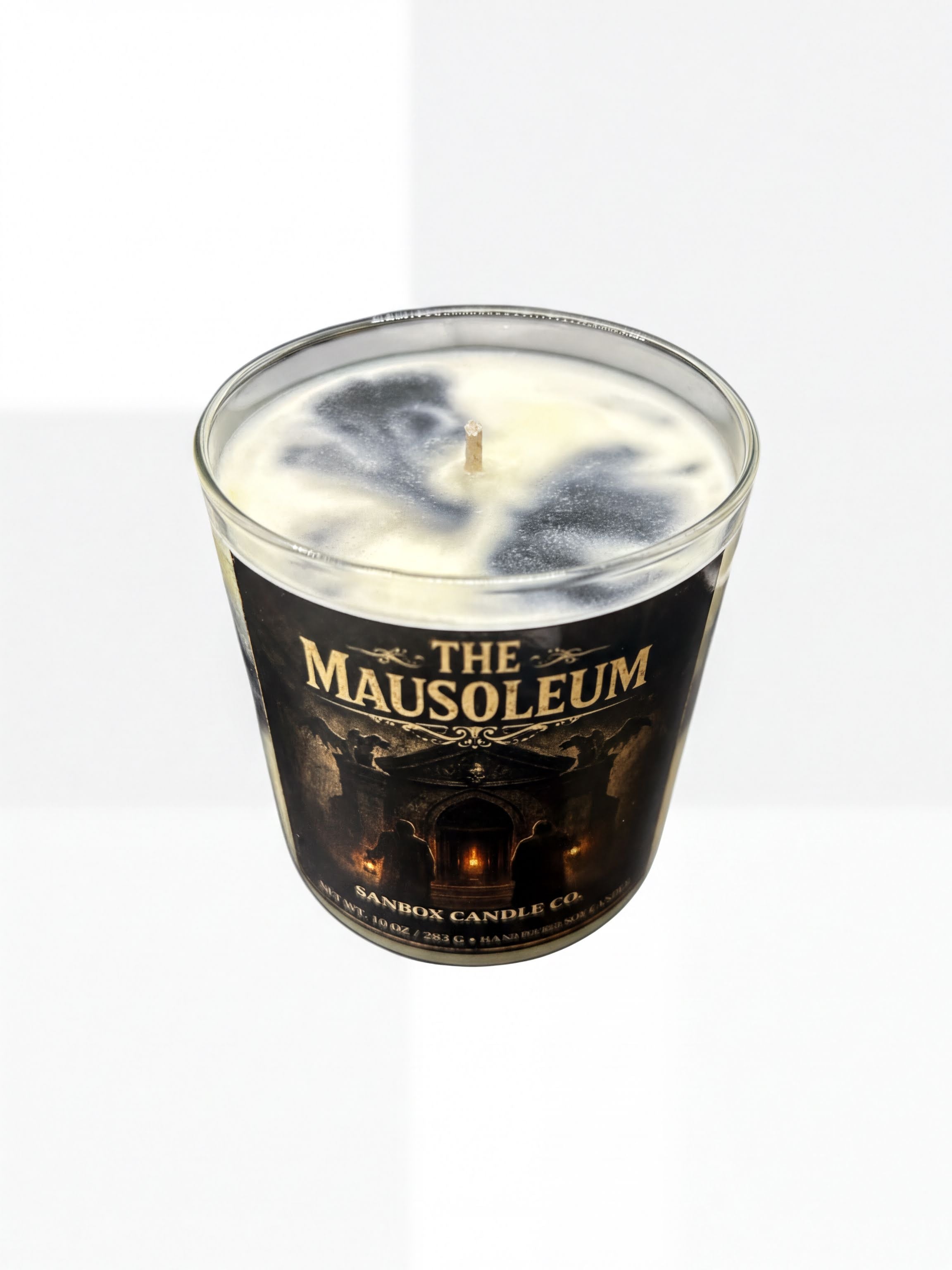A glass candle holder with a candle inside, featuring a label that reads "The Mausoleum" and "SANBOX CANDLE CO."