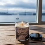 A lit candle in a glass jar sits on a wooden surface, with a view of a bridge and water in the background through a window.