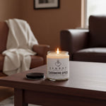A lit candle in a glass jar sits on a wooden table, with a brown leather couch and a framed picture in the background.