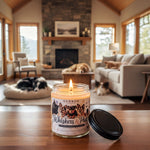 A lit candle in a jar with a label that reads "Whiskers & Paws" sits on a wooden table in a cozy living room with a fireplace, a dog sleeping on a bed, and a cat sitting on a couch.
