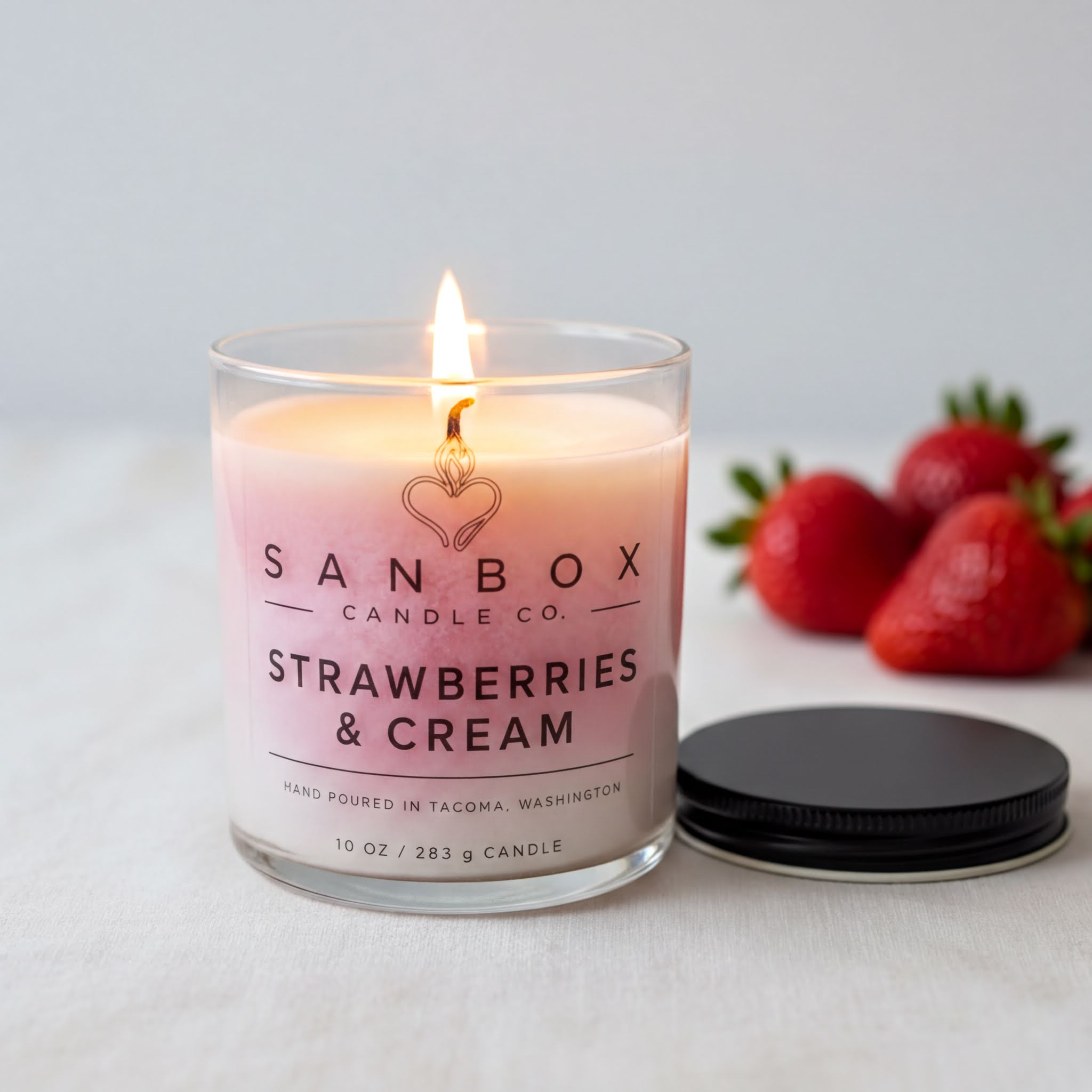A lit candle in a glass container with a lit flame, surrounded by fresh strawberries on a white surface.