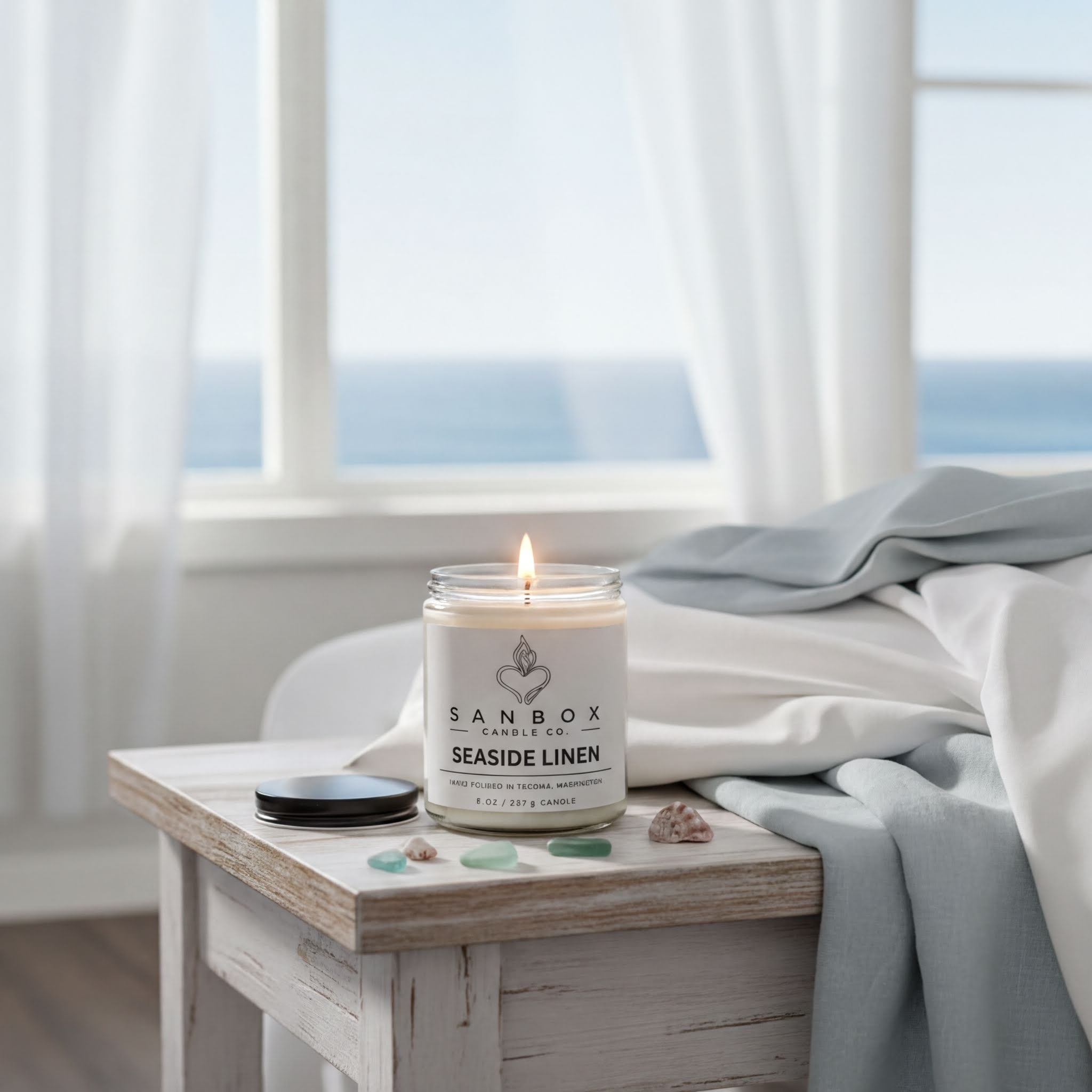 A lit candle in a jar sits on a wooden table, with a view of the ocean visible through a window in the background.