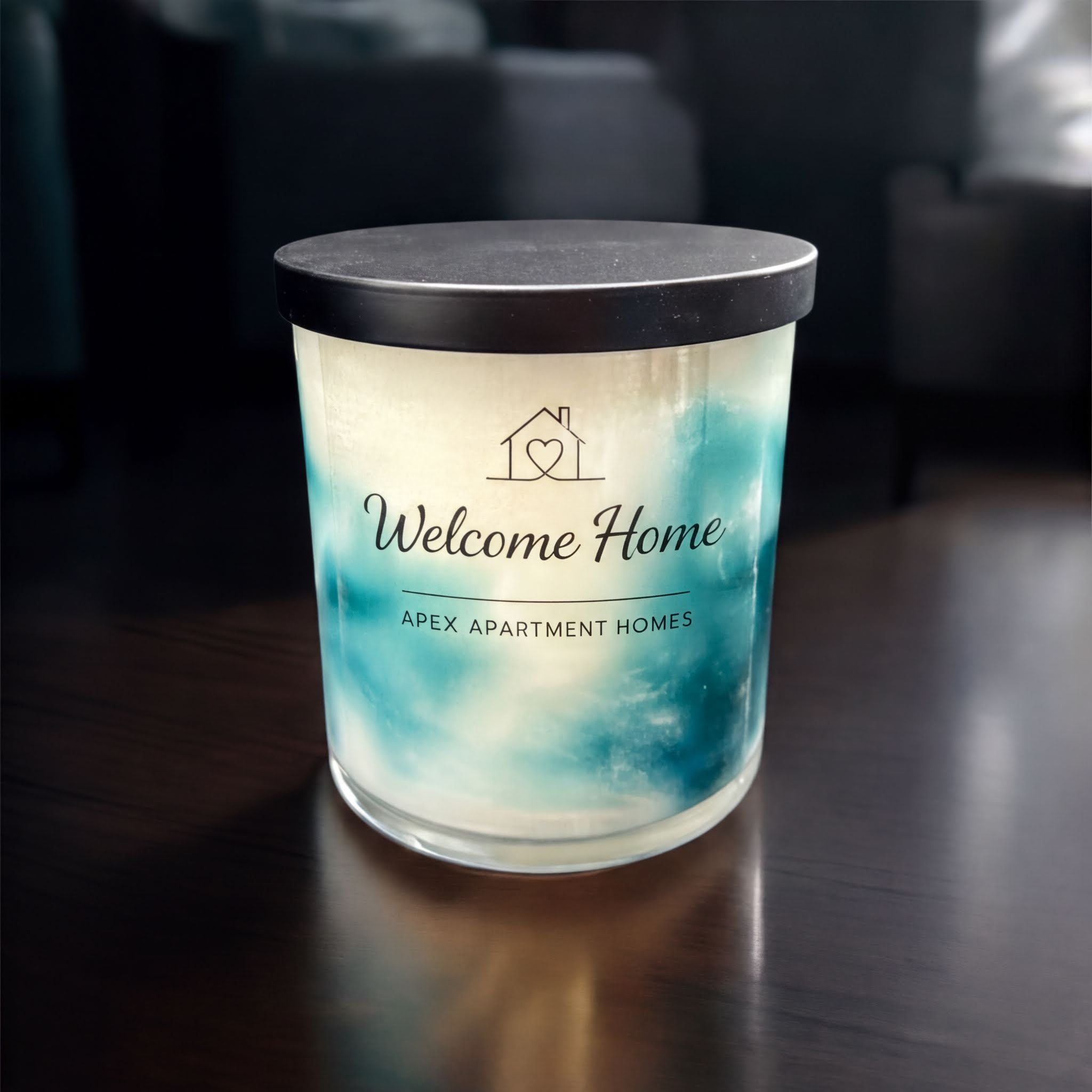 A candle labeled "Welcome Home" sits on a wooden surface, with a blurred background that suggests a cozy indoor setting.