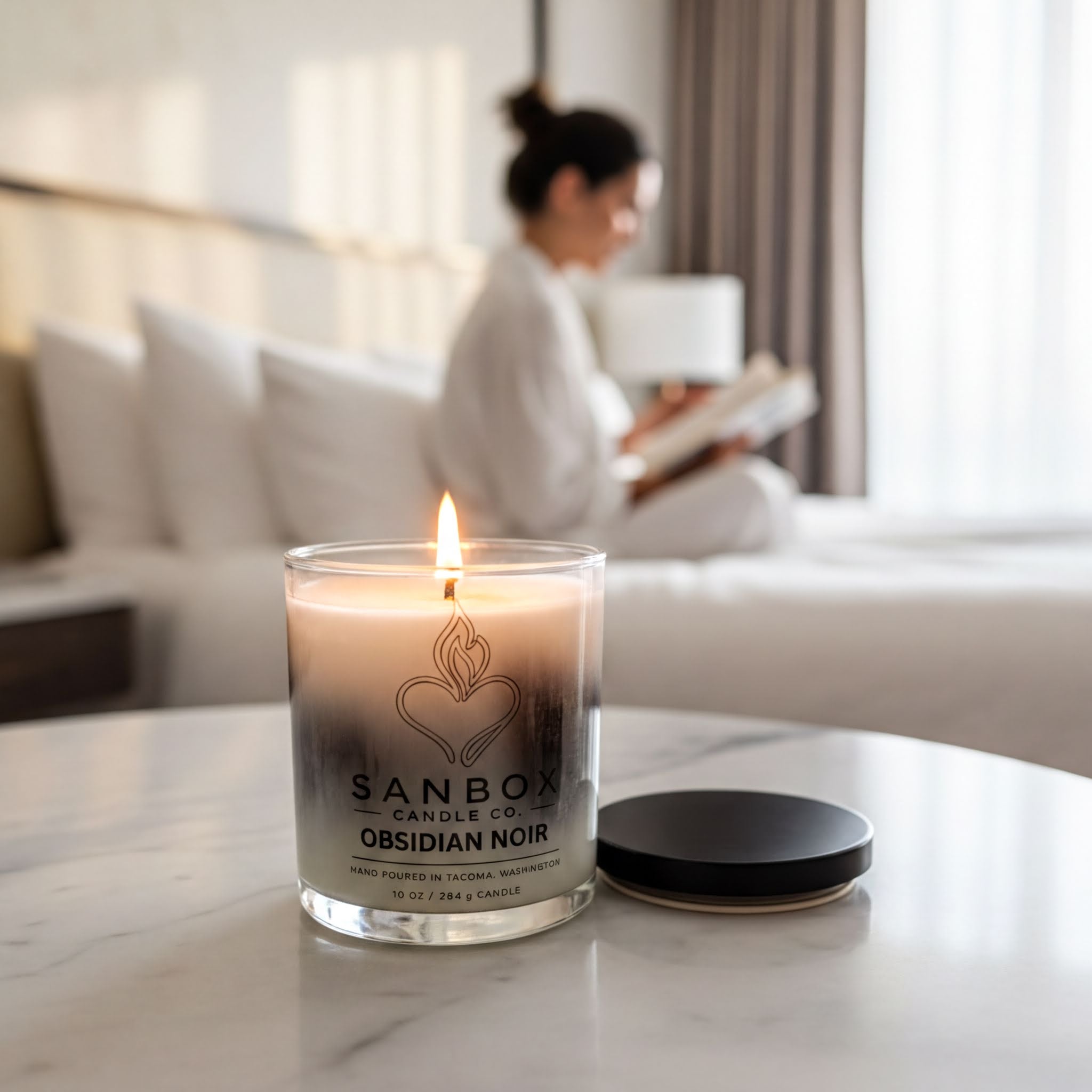 A lit candle in a glass container sits on a marble surface, with a blurred image of a person sitting on a bed in the background.
