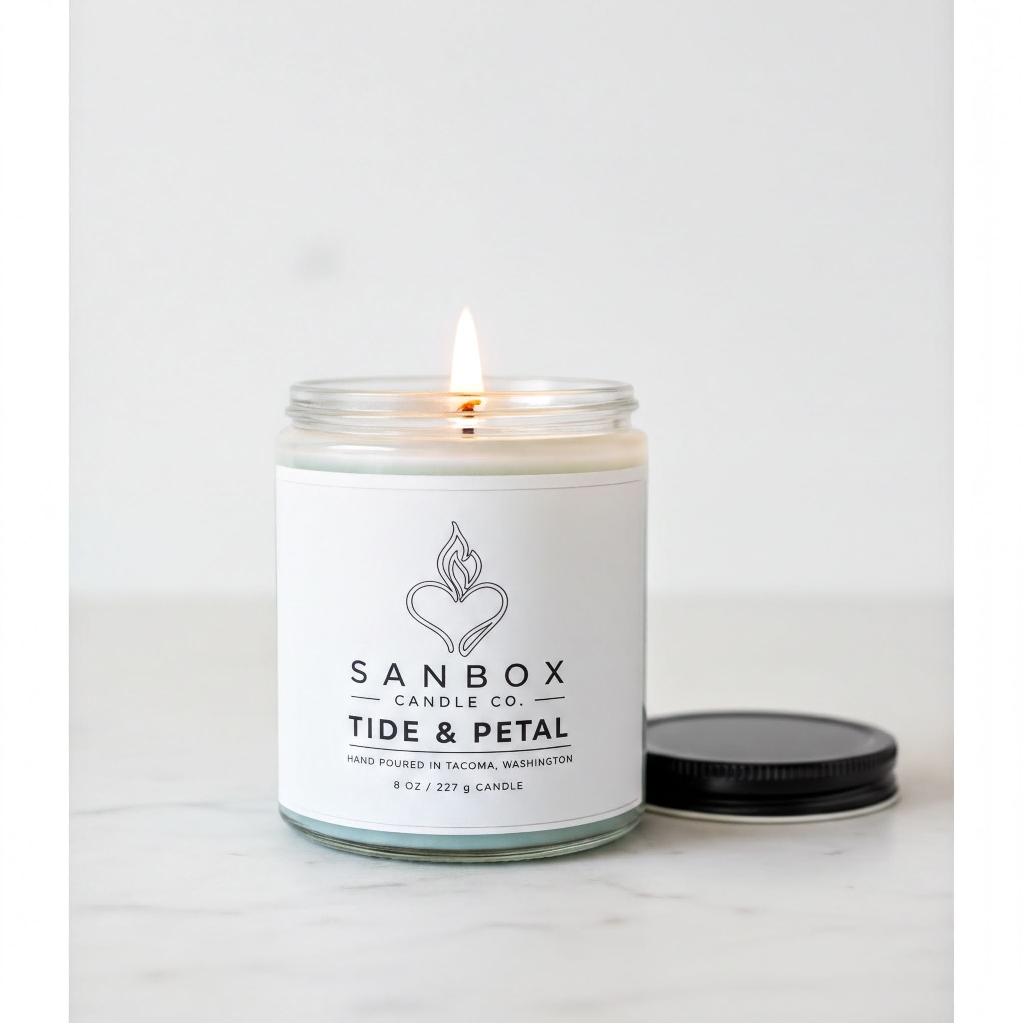 A white candle with a lit flame sits on a white surface, accompanied by a black lid.