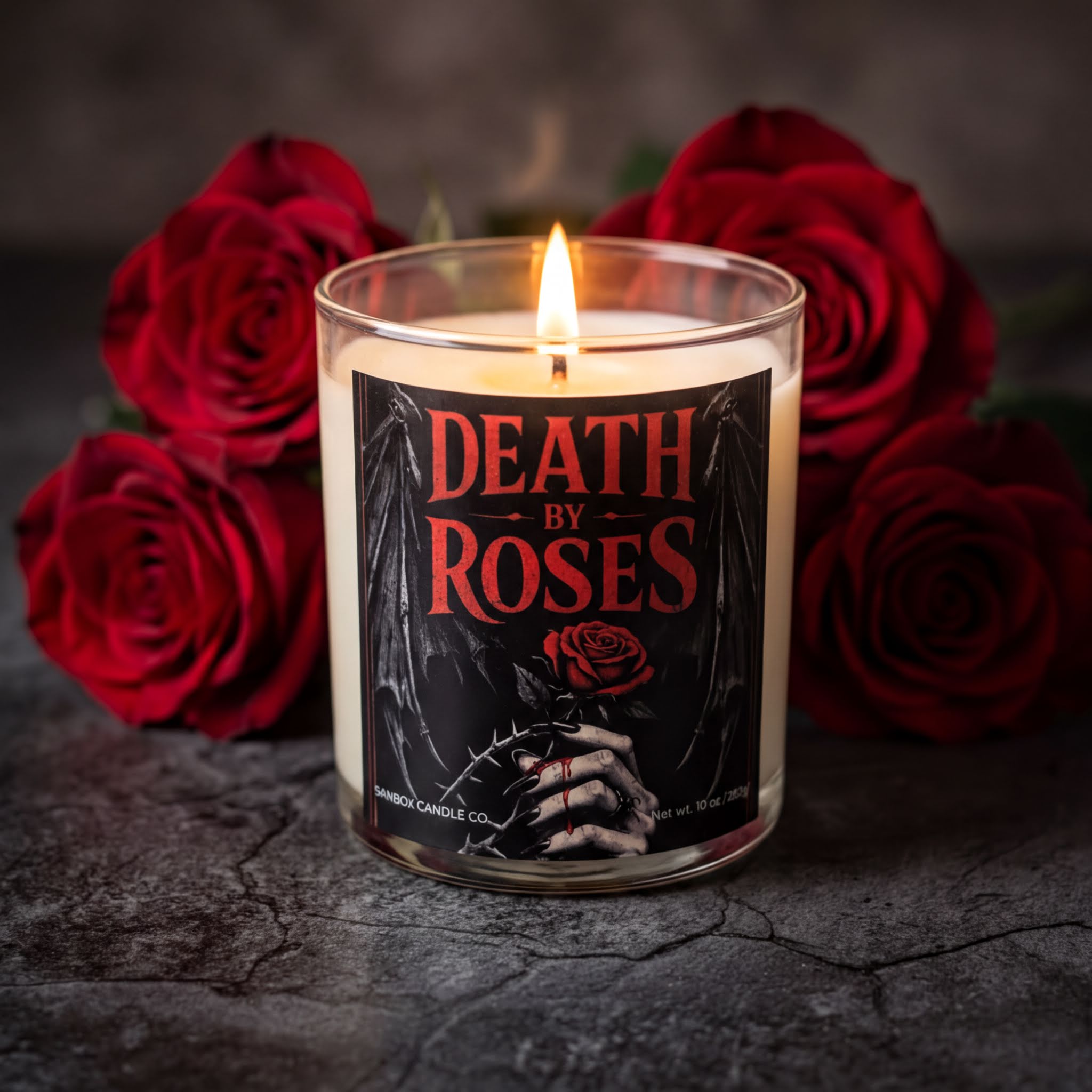 A lit candle with the words "DEATH BY ROSES" on it, surrounded by red roses, on a dark surface.