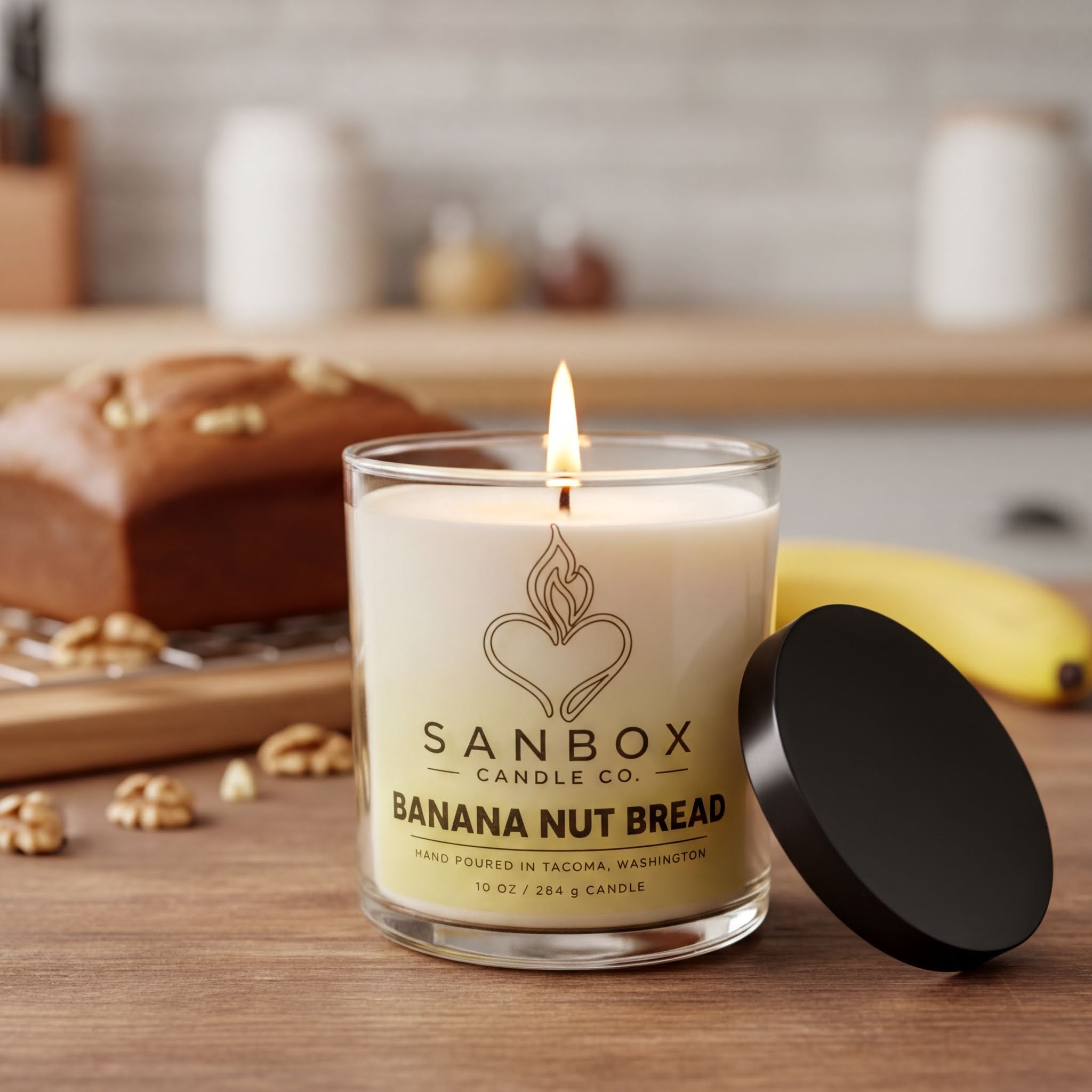 A lit candle in a glass container labeled "SANBOX CANDLE CO. BANANA NUT BREAD" sits on a wooden surface, with a loaf of banana nut bread and some nuts in the background.
