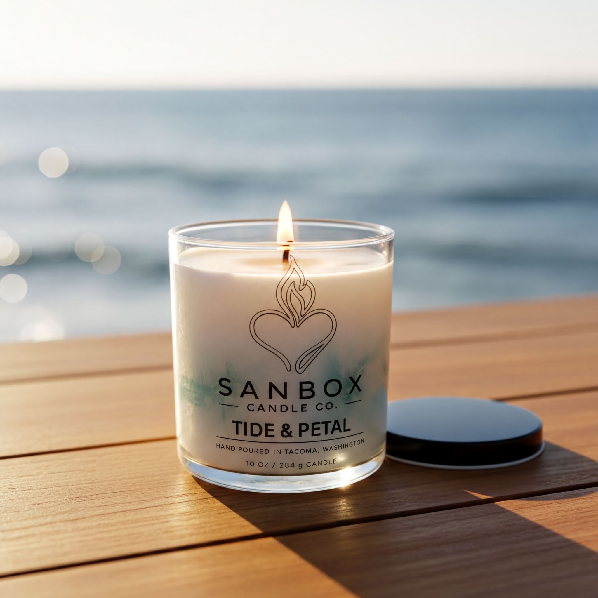 A lit candle in a glass container sits on a wooden surface, with the ocean visible in the background.