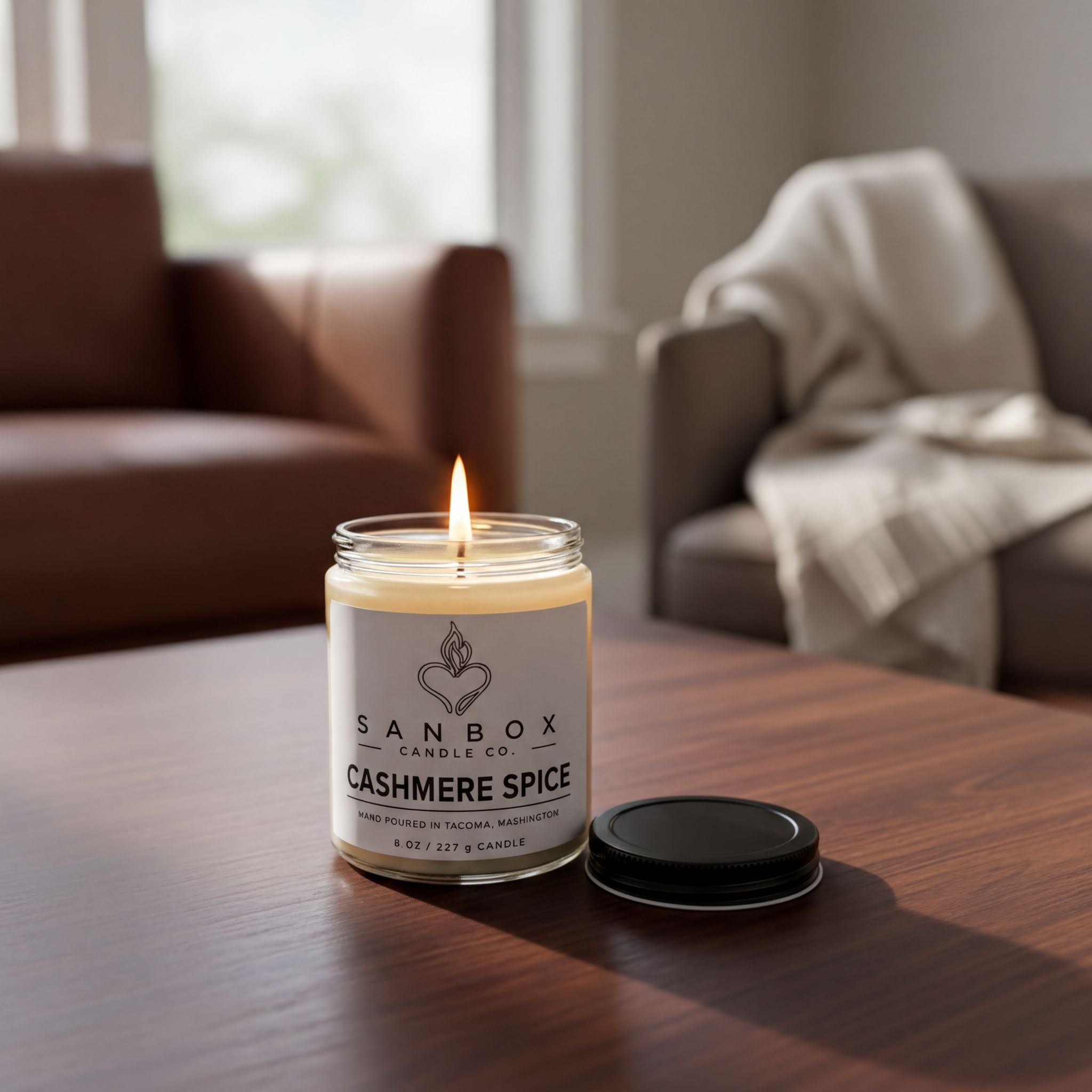 A lit candle in a glass jar labeled "SANBOX CANDLE CO. CASHMERE SPICE" sits on a wooden table, with a cozy blanket draped over a chair in the background.