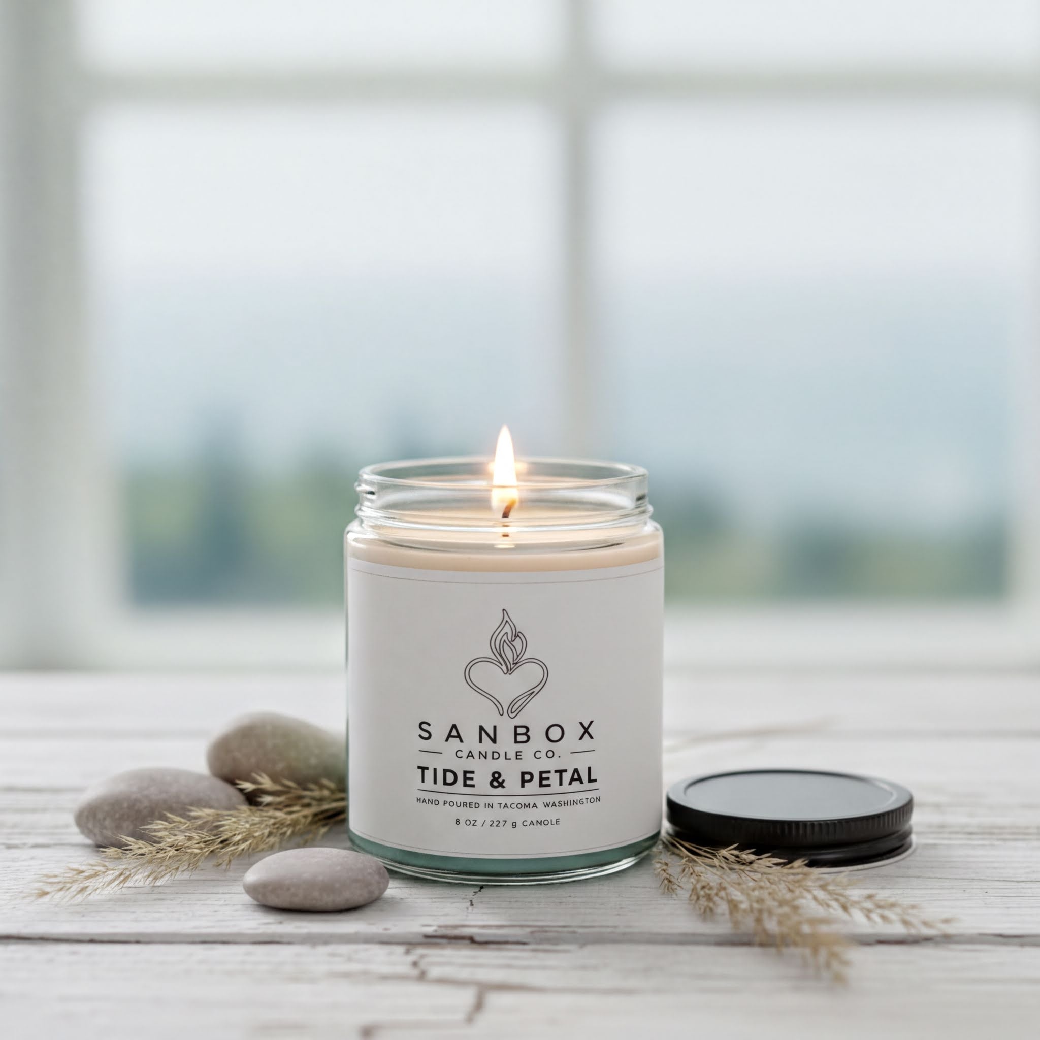 A lit candle in a glass jar with a label that reads "SANBOX Candle Co. TIDE & PETAL" sits on a wooden surface, surrounded by some rocks and dried plants.