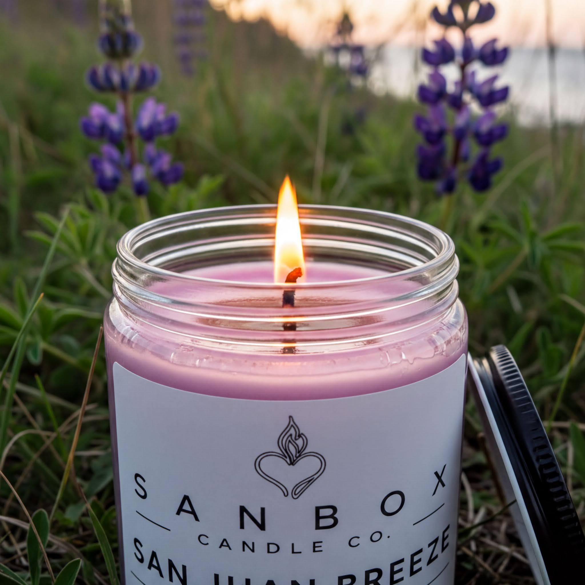 A lit pink candle in a jar, with a label that reads "SANBOX CANDLE CO. SAN JUAN BREZE", set against a backdrop of purple flowers and a grassy field.