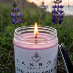 A lit pink candle in a jar, with a label that reads "SANBOX CANDLE CO. SAN JUAN BREZE", set against a backdrop of purple flowers and a grassy field.