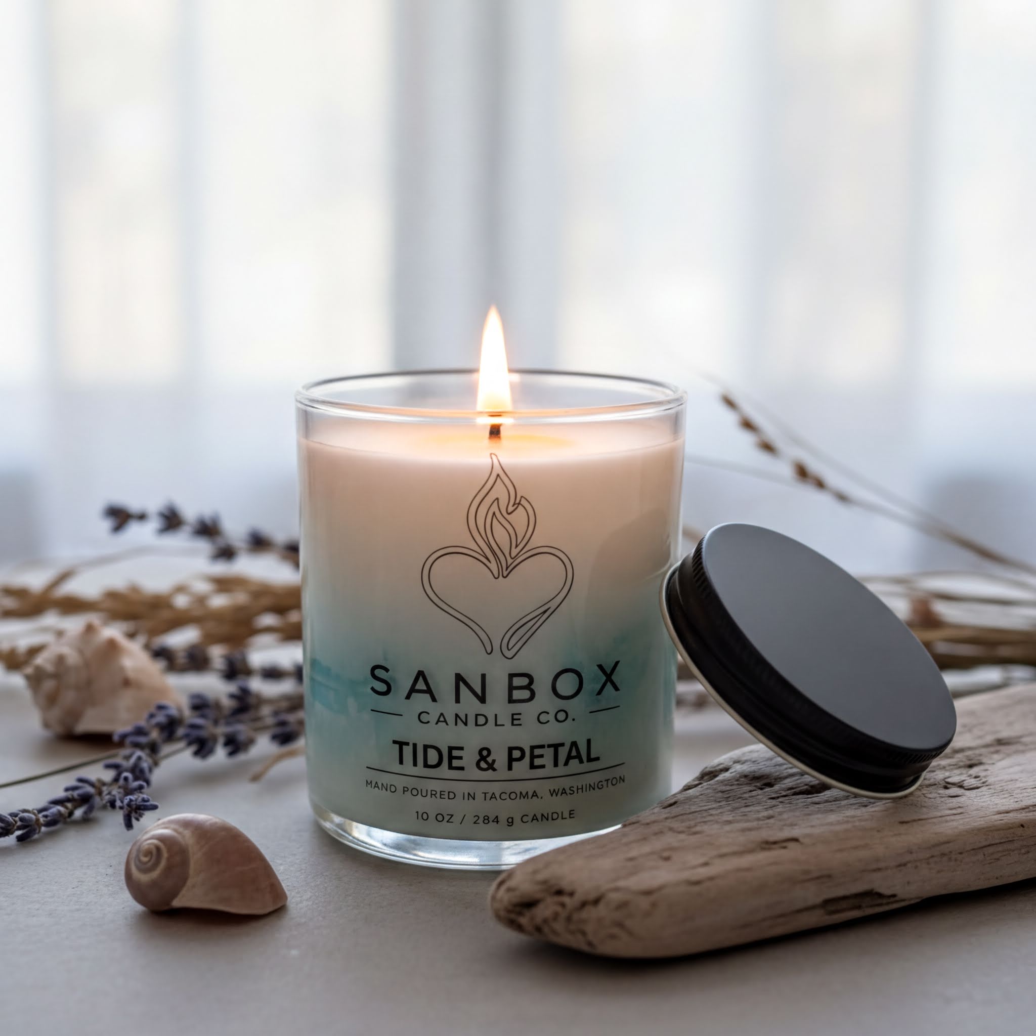 A lit candle in a glass container with a black lid, surrounded by seashells, dried lavender, and driftwood on a table.