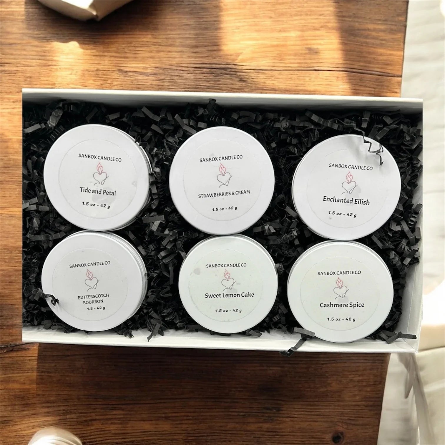 Box of six scented candle sample tins in assorted fragrances on a wooden table.