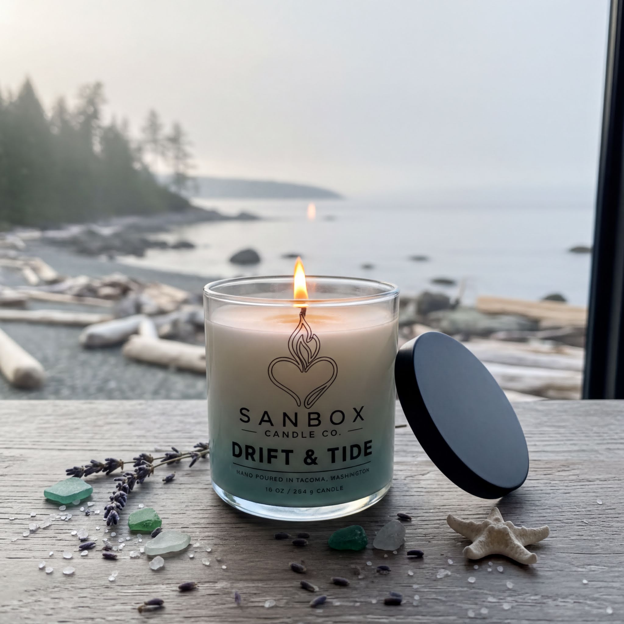 A lit candle in a glass container sits on a wooden surface, with a blurred background of a beach and trees.