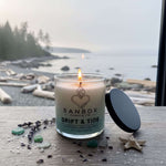 A lit candle in a glass container sits on a wooden surface, with a blurred background of a beach and trees.