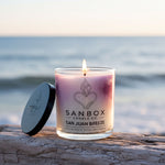 A lit candle in a glass container sits on a wooden surface, with a blurred ocean and sky in the background.