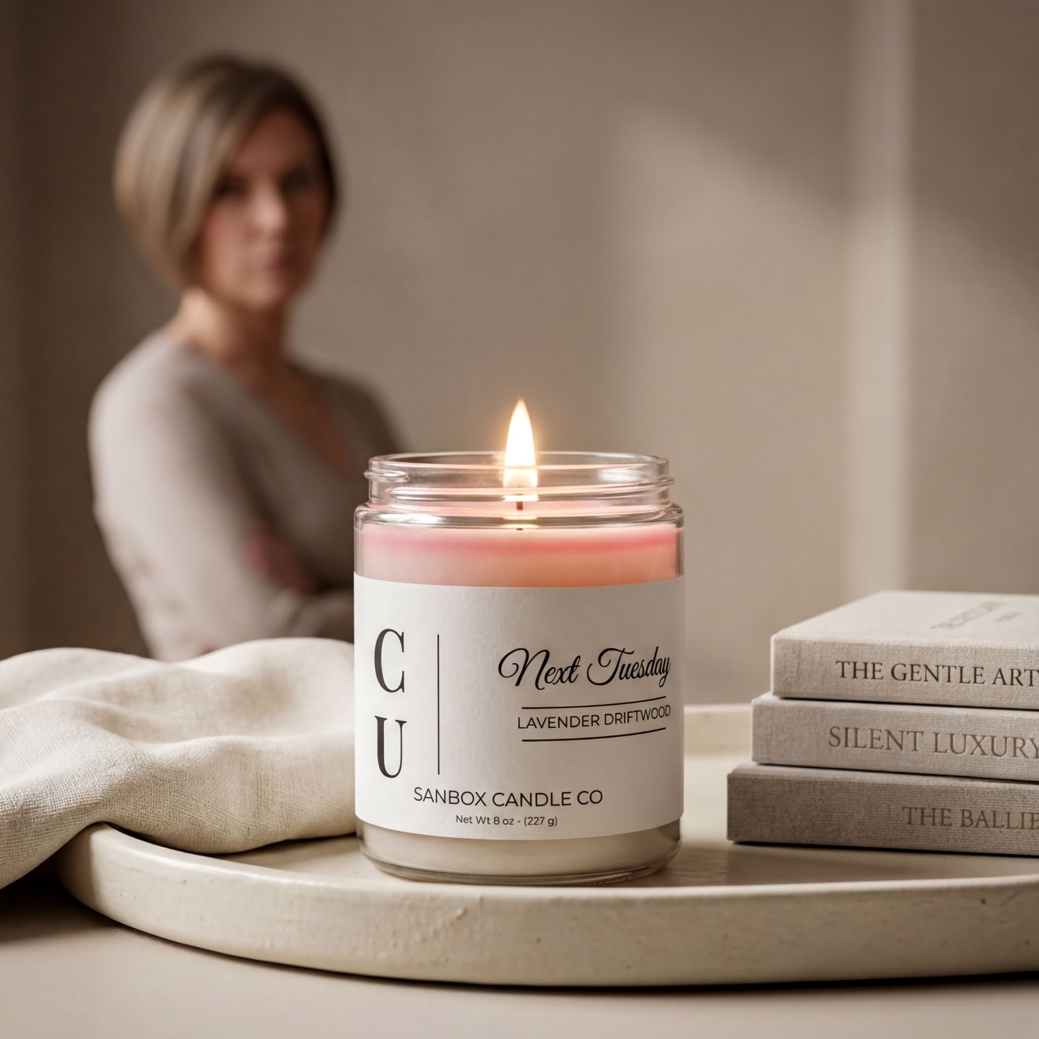 a lit candle in a jar, with a blurred woman in the background. The candle is labeled "Next Tuesday Lavender Driftwood" and is placed on a white tray. There are also some books visible in the background.