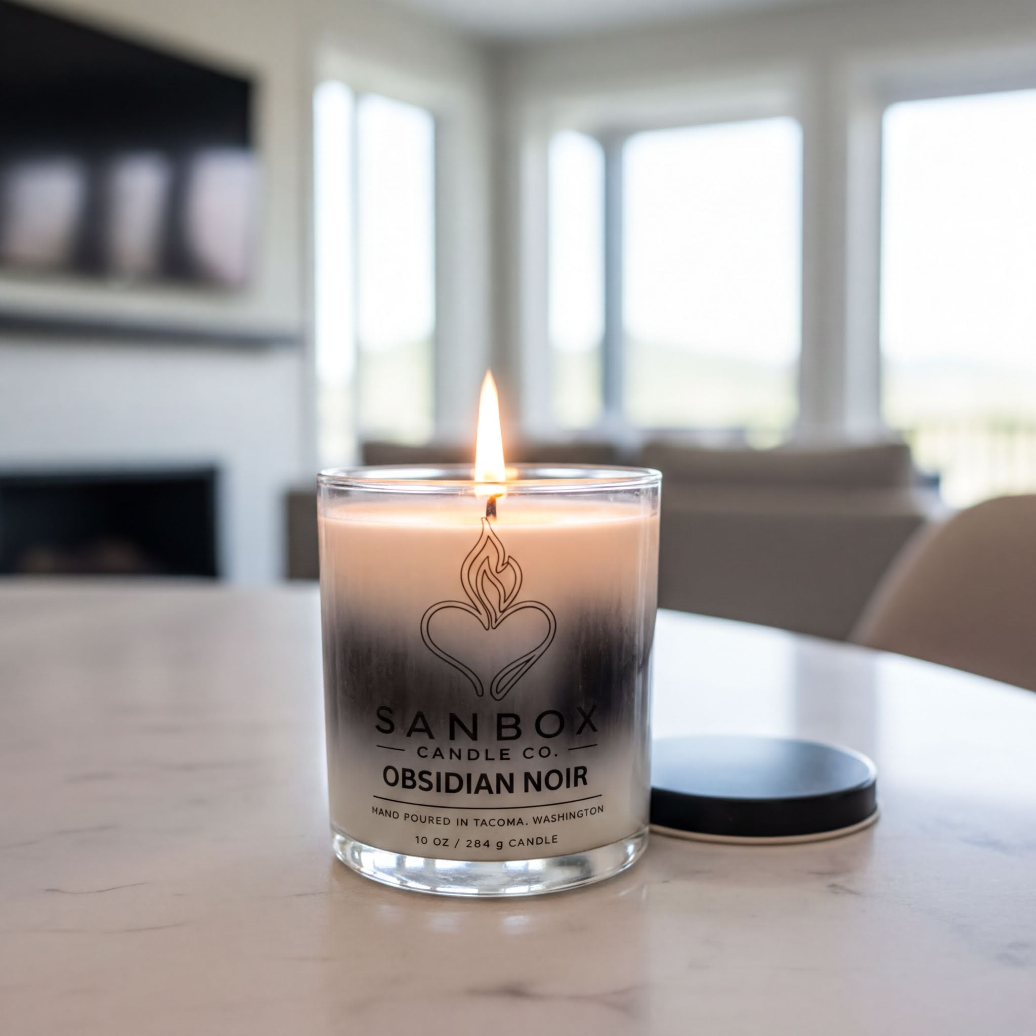 A lit candle in a glass container with a label that reads "SANBBOX CANDLE CO. OBSIDIAN NOIR" sits on a marble countertop.