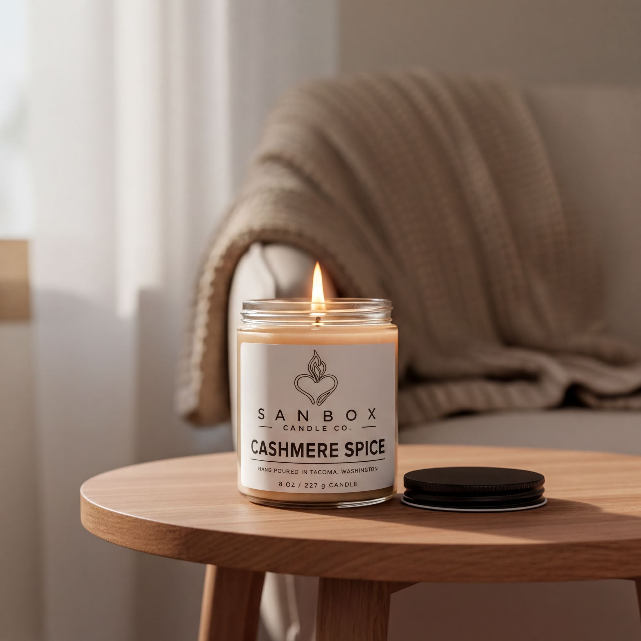 A lit candle in a glass jar labeled "SANBOX CANDLE CO. CASHMERE SPICE" sits on a wooden table, with a beige blanket draped over a chair in the background.