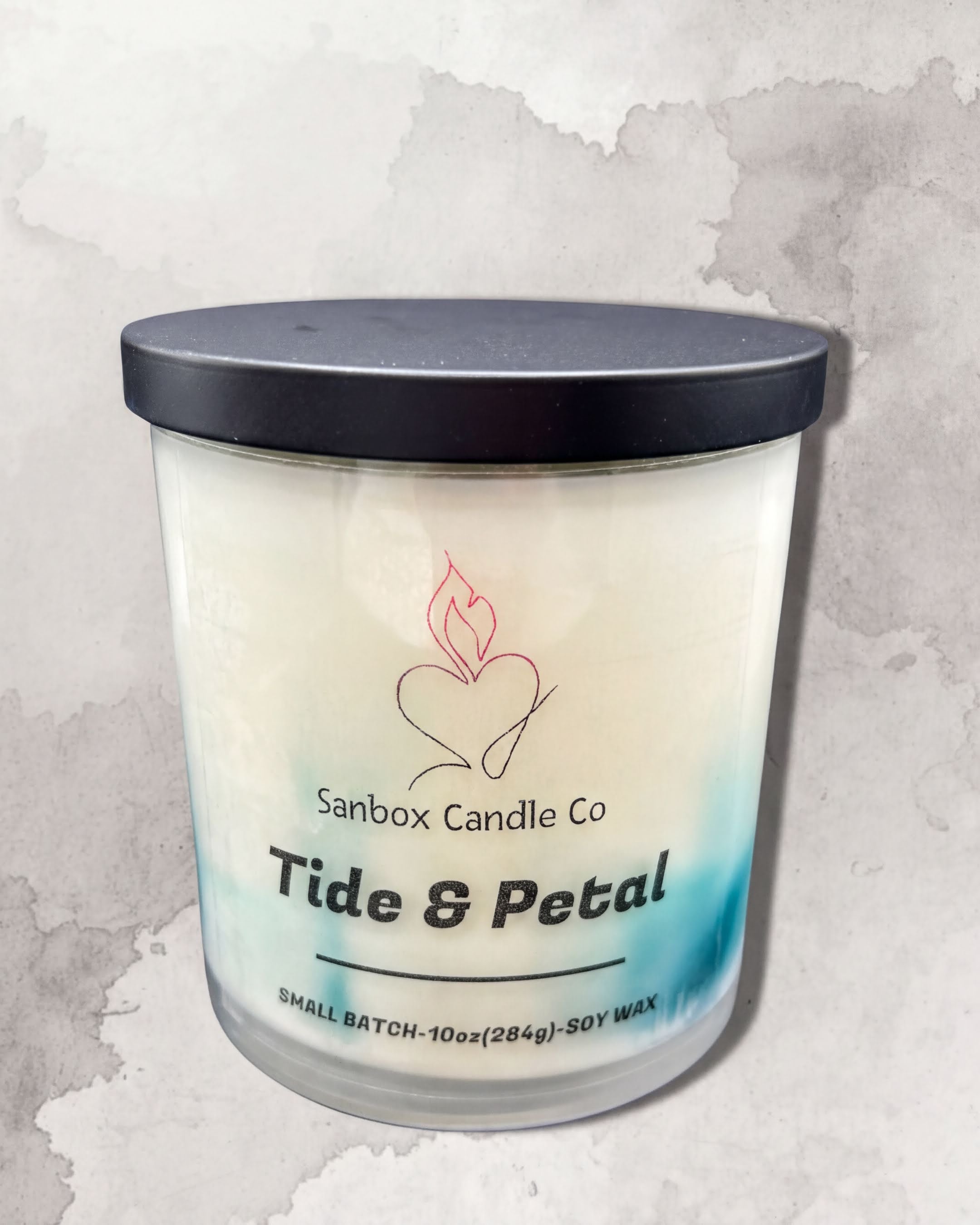 a candle in a cylindrical container with a black lid, placed against a textured gray background.
