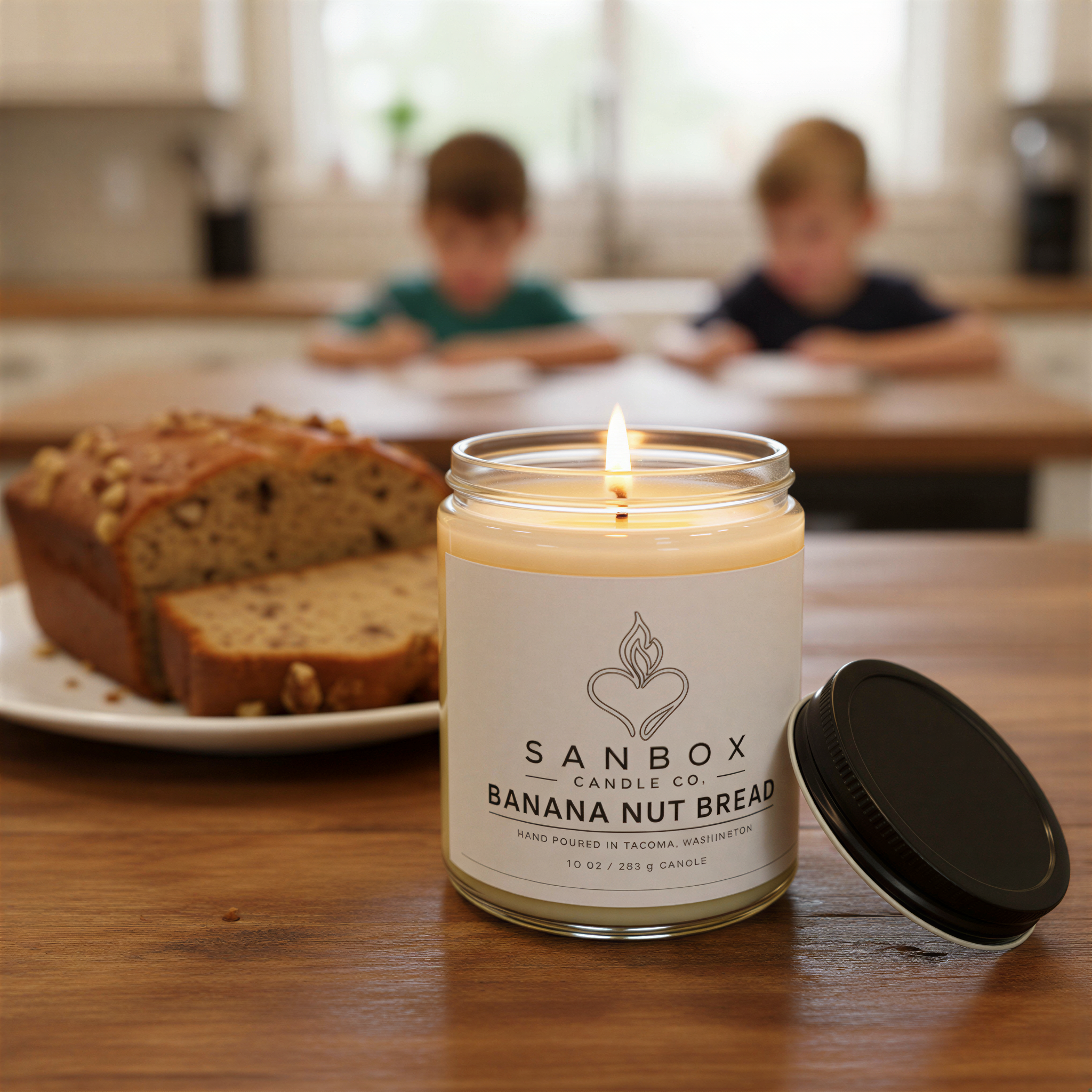 A candle labeled "SANBOX BANANA NUT BREAD" sits on a wooden table, with a slice of banana nut bread in the background.