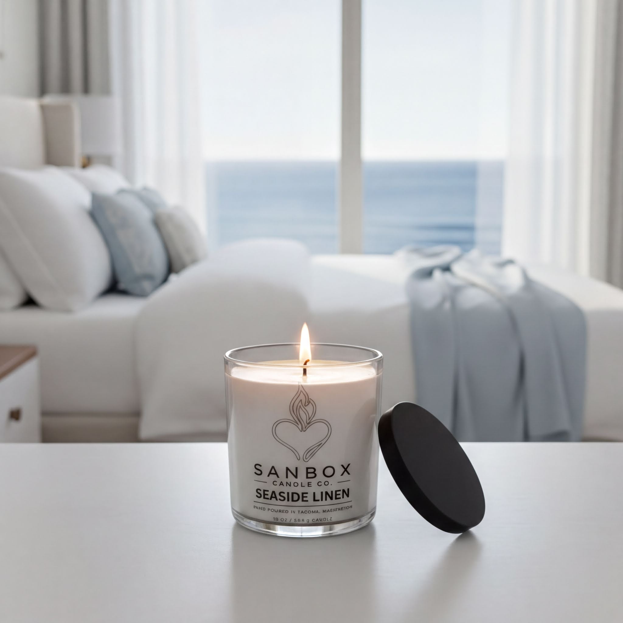 A lit candle in a glass container sits on a white surface, with a view of a window overlooking the ocean in the background.
