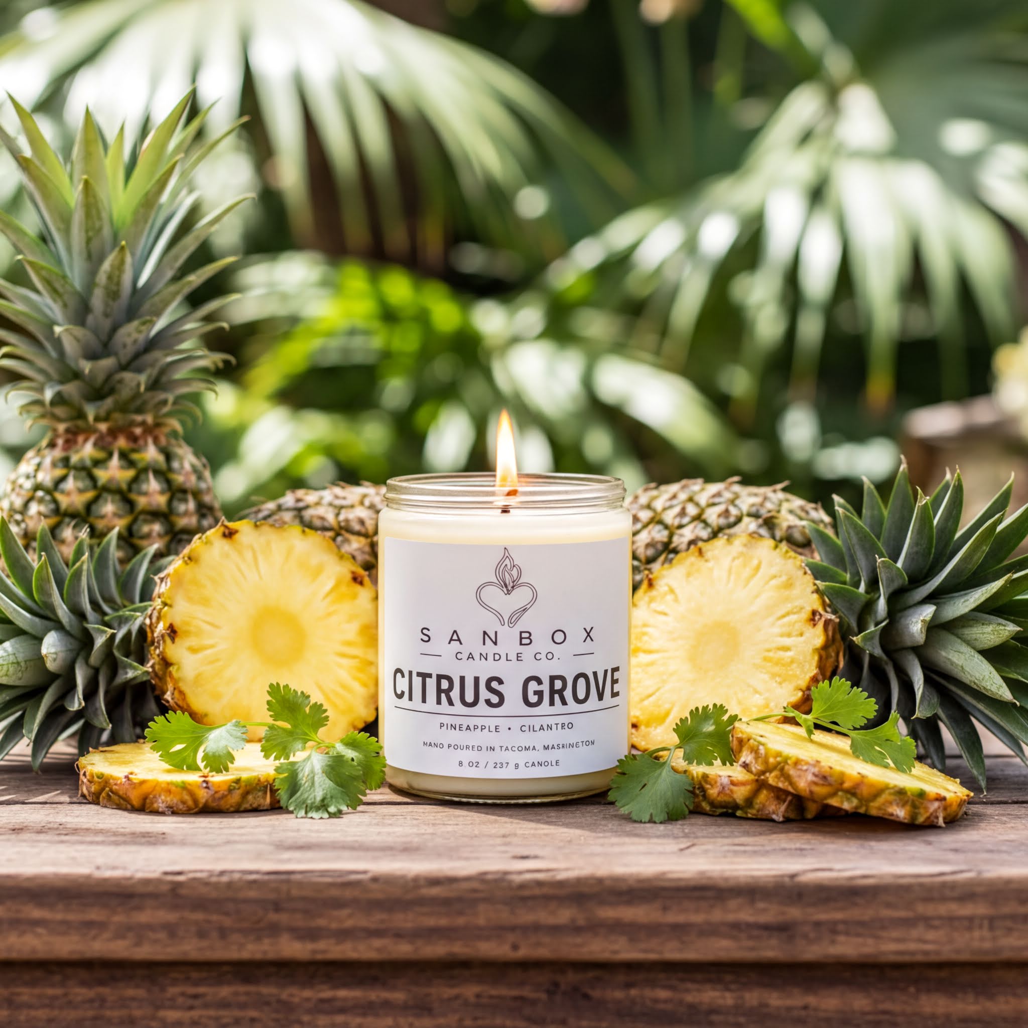 A lit candle labeled "Citrus Grove" surrounded by sliced pineapples and cilantro on a wooden surface, with a blurred background of tropical foliage.