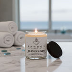 A lit candle in a glass jar labeled "SANBOX CANDLE CO. SEASIDE LINEN" sits on a marble countertop, with rolled towels and a window in the background.
