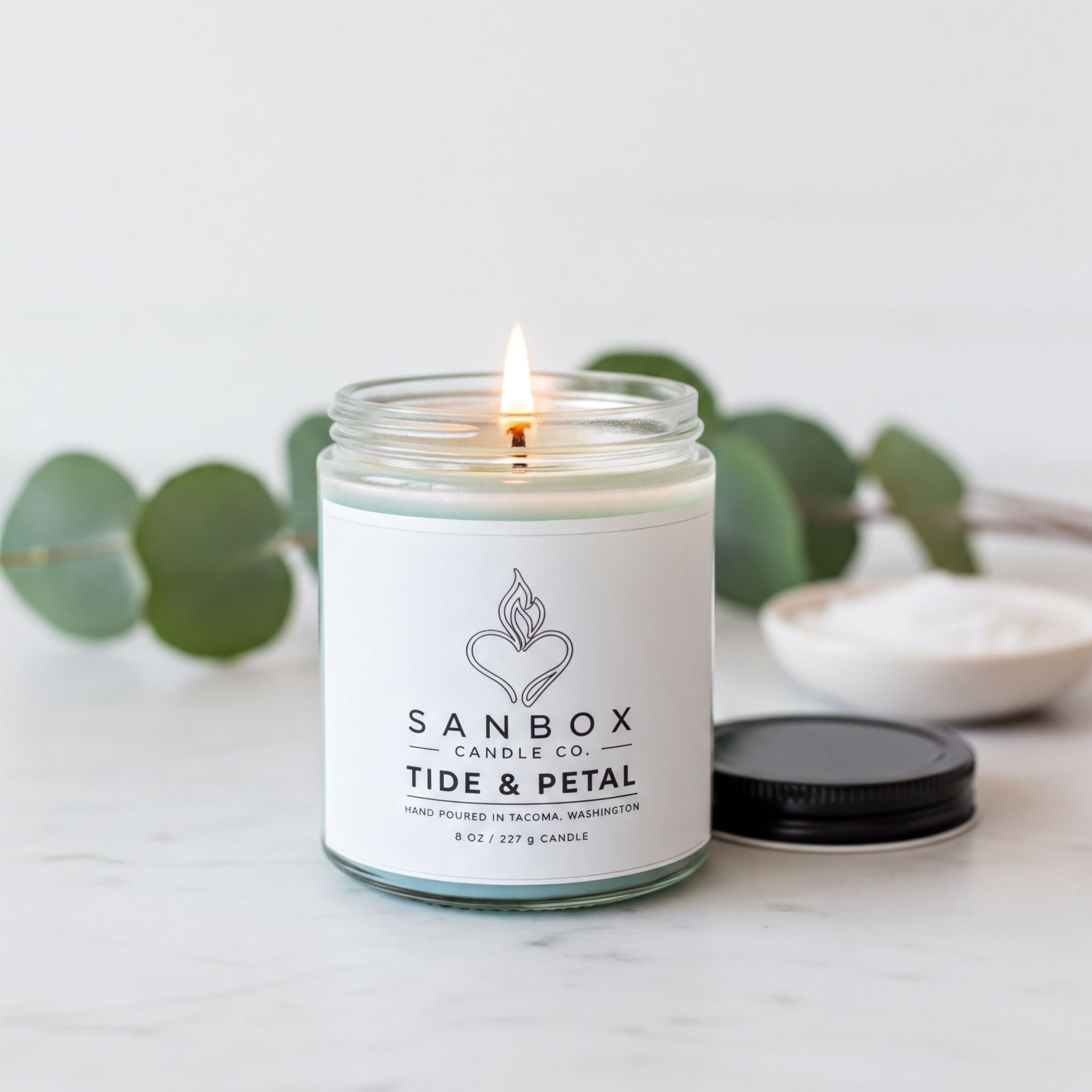 A lit candle in a glass jar with a white label, surrounded by green leaves and a small white bowl, on a white marble surface.