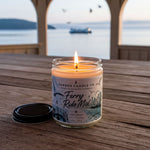 A lit candle in a jar sits on a wooden table, with a scenic view of a body of water and boats in the background.