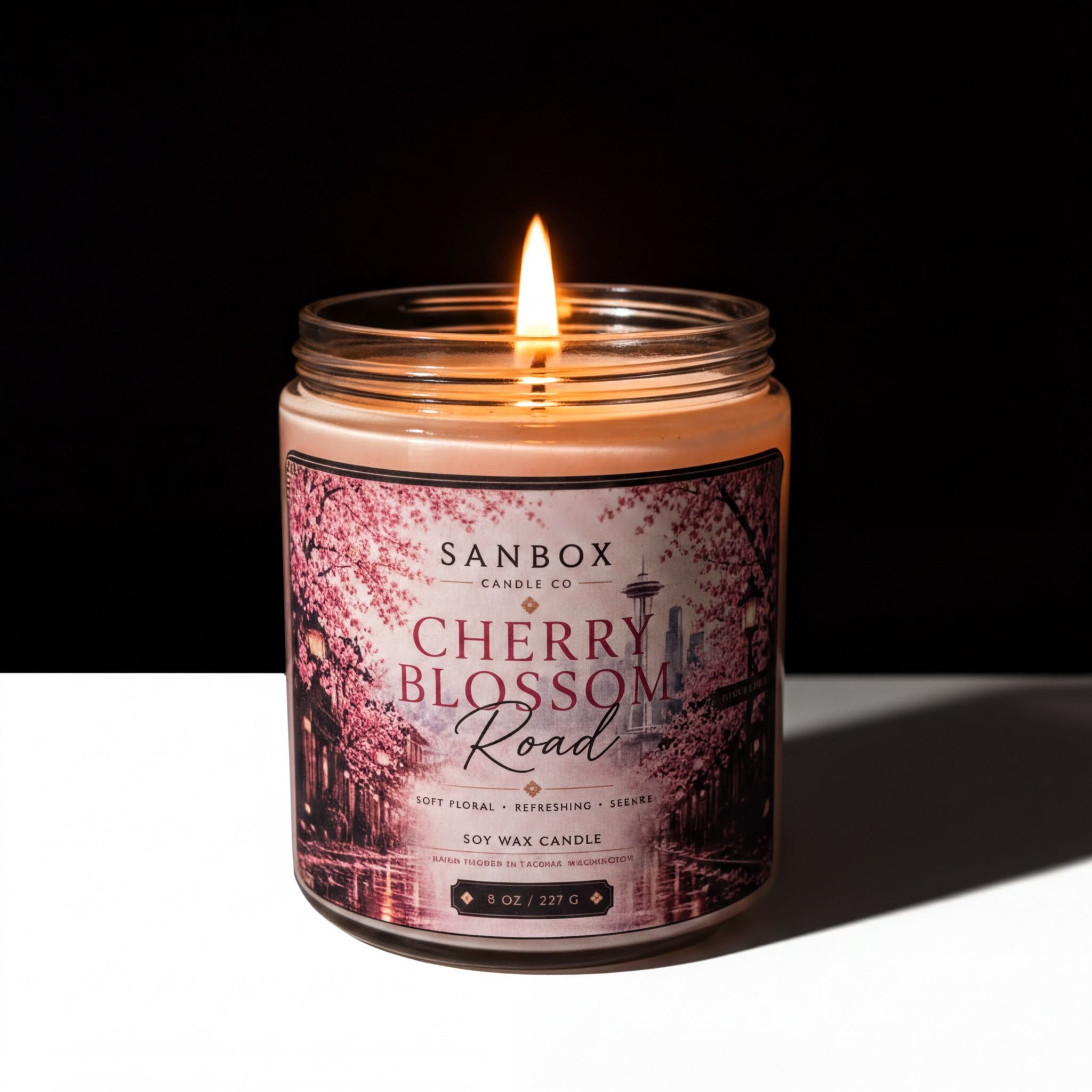 A lit soy wax candle in a jar with a label that reads "SANBOX Candle Co Cherry Blossom Road" and "Soft Floral - Refreshing - Scented".