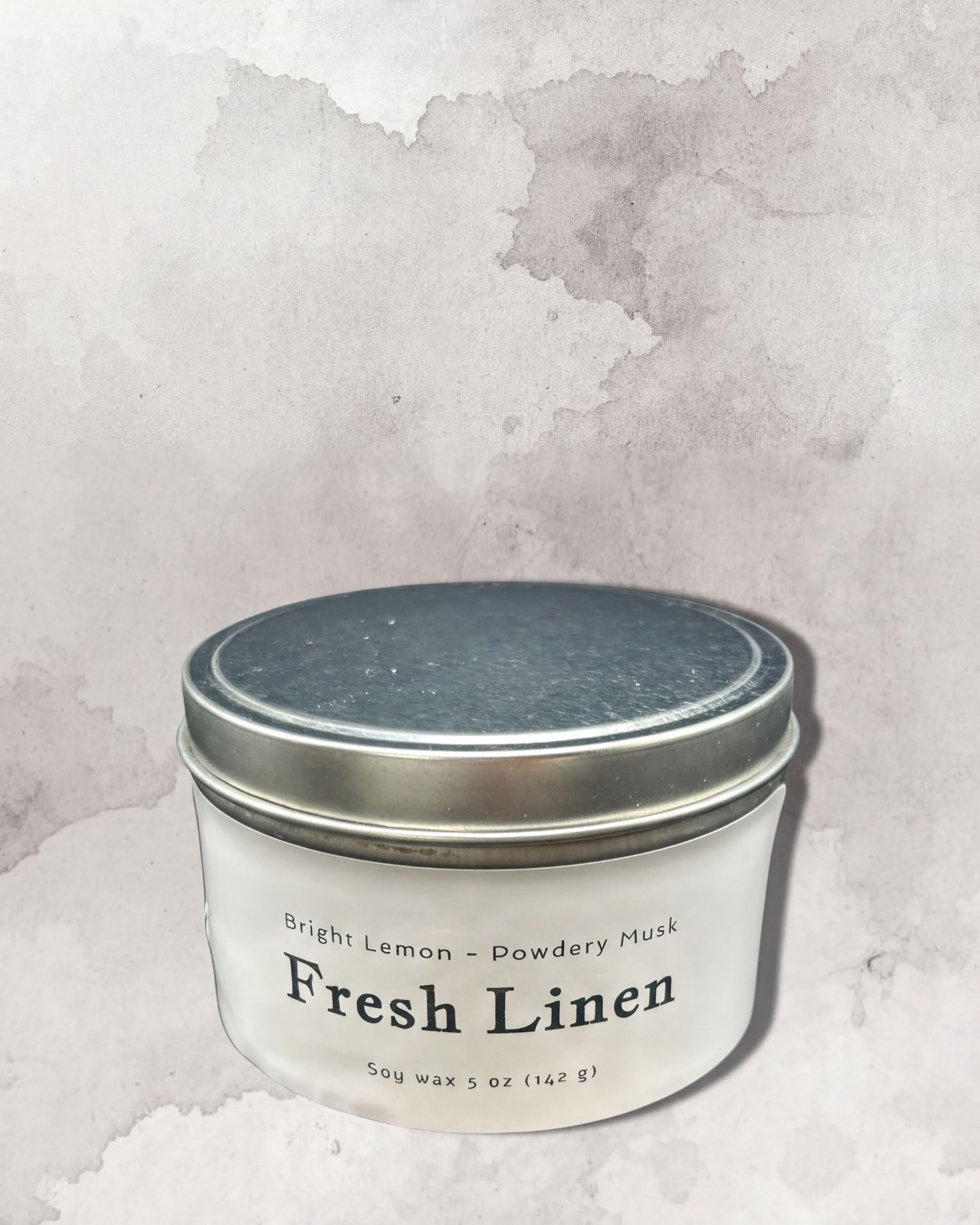 A white tin candle labeled "Fresh Linen" with a silver lid, placed against a textured gray background.