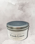 A white tin candle labeled "Fresh Linen" with a silver lid, placed against a textured gray background.