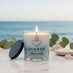 A lit candle in a glass jar with a black lid, placed on a marble surface, with a blurred background of the ocean and greenery.