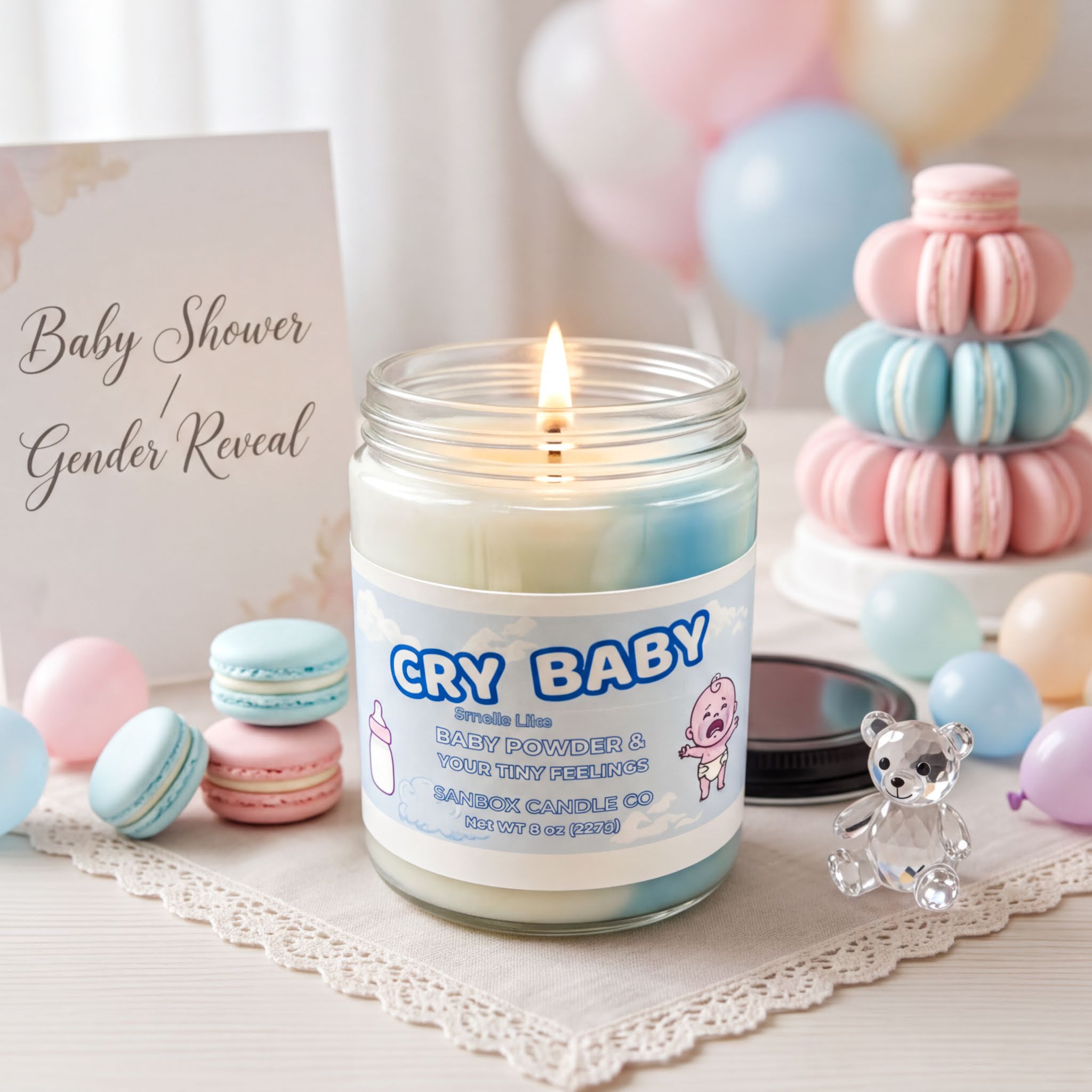 a candle labeled "CRY BABY" surrounded by various baby-related items, including macarons, balloons, and a teddy bear.